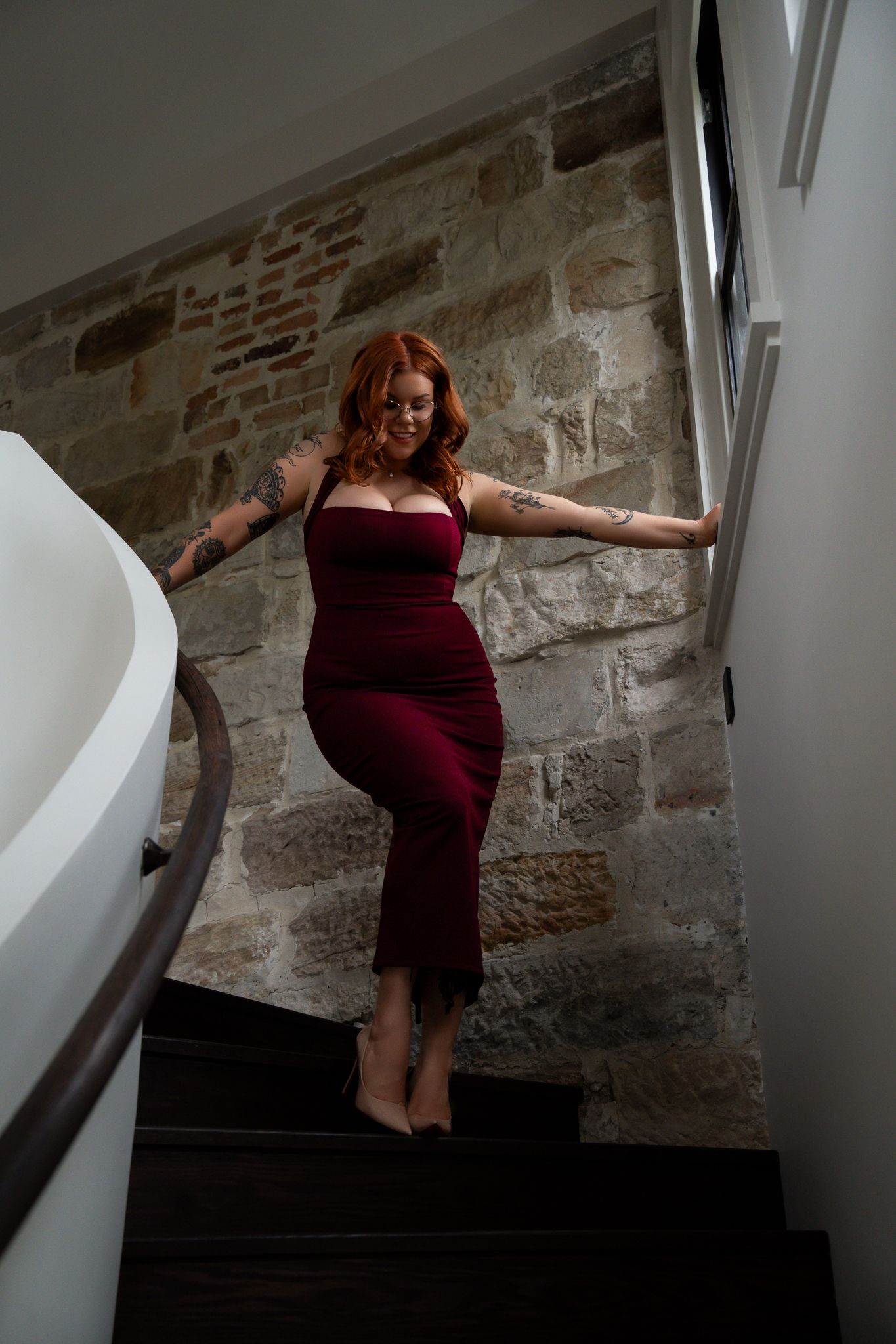 A curvy redhead woman with tattoos poses in a burgundy midi dress on stone-walled stairs.