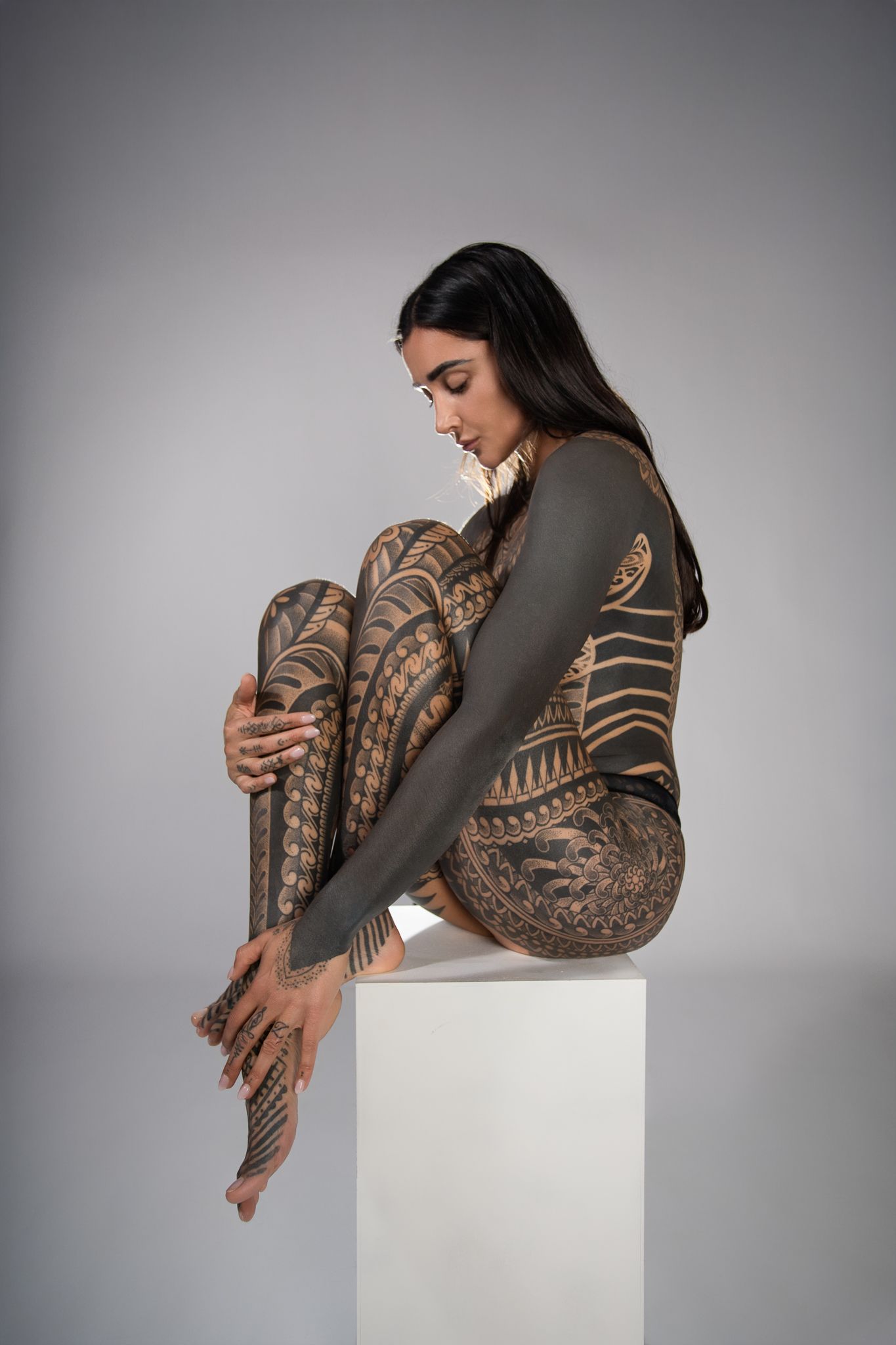 A person with intricate tribal tattoo sleeves poses artistically on a white pedestal against a gray background.