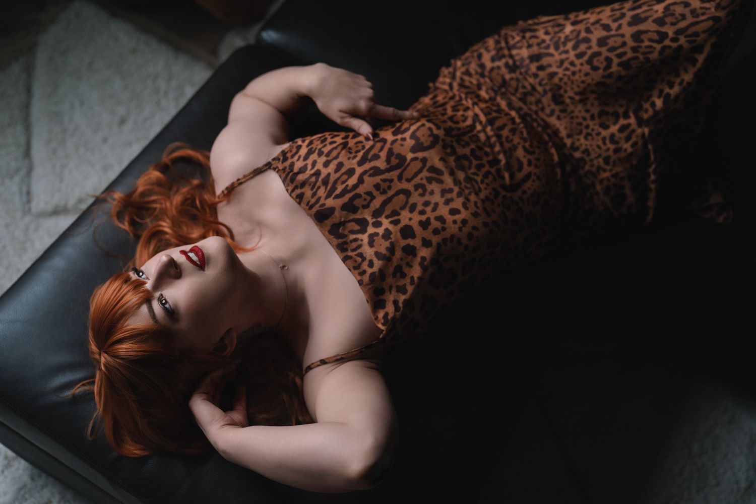 Person with red hair wearing a leopard print dress lying on dark leather furniture.