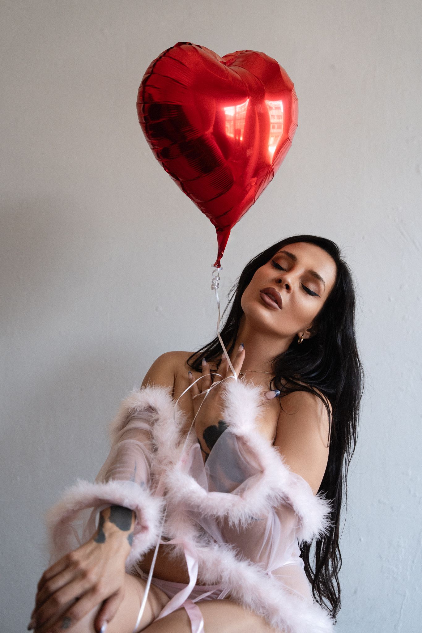 Woman in lingerie and feather robe holding a red heart-shaped balloon for Valentine's Day.