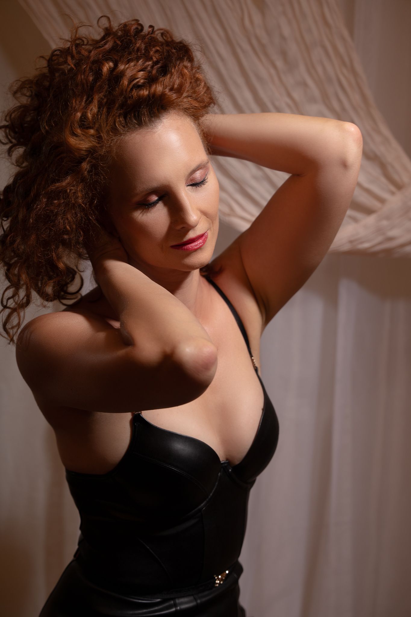 A woman with curly red hair poses in a black leather corset top with hands behind her head.