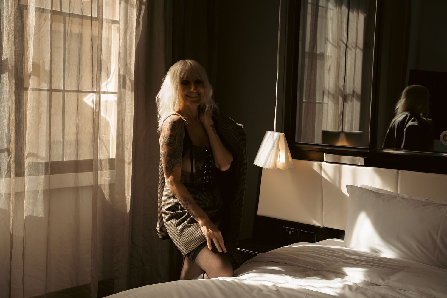 A blonde woman with tattoos sits on a hotel bed in moody natural light near sheer curtains.