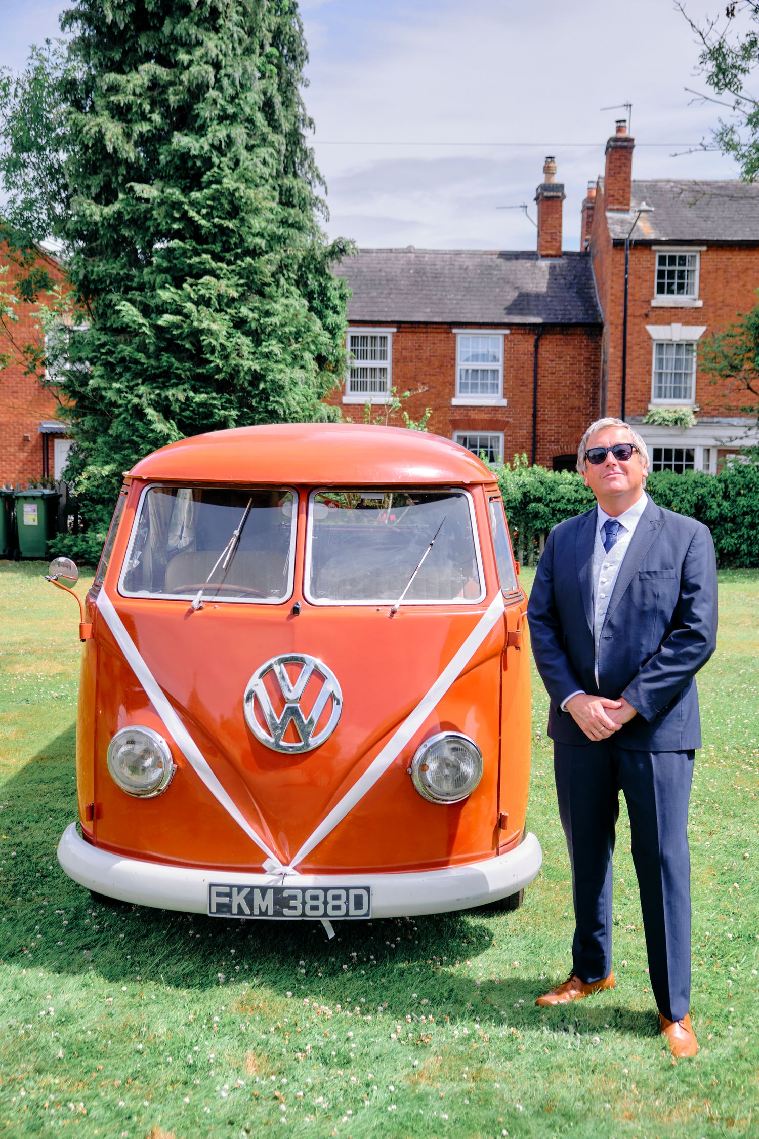 Man in navy suit poses beside a vintage orange VW camper van decorated with white ribbons outdoors.