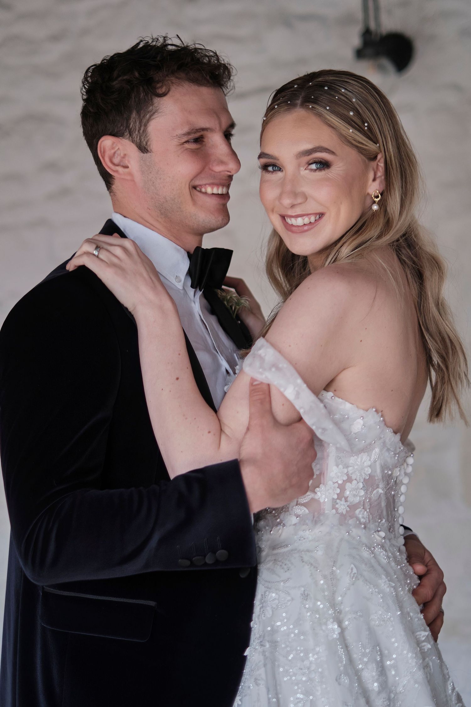 Smiling bride in off-shoulder lace wedding dress poses with groom in dark suit against white brick wall.