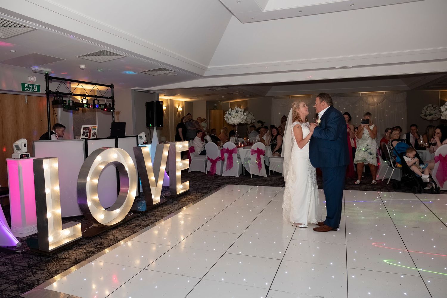 Bride and groom share first dance beside illuminated LOVE letters on a white wedding dance floor.