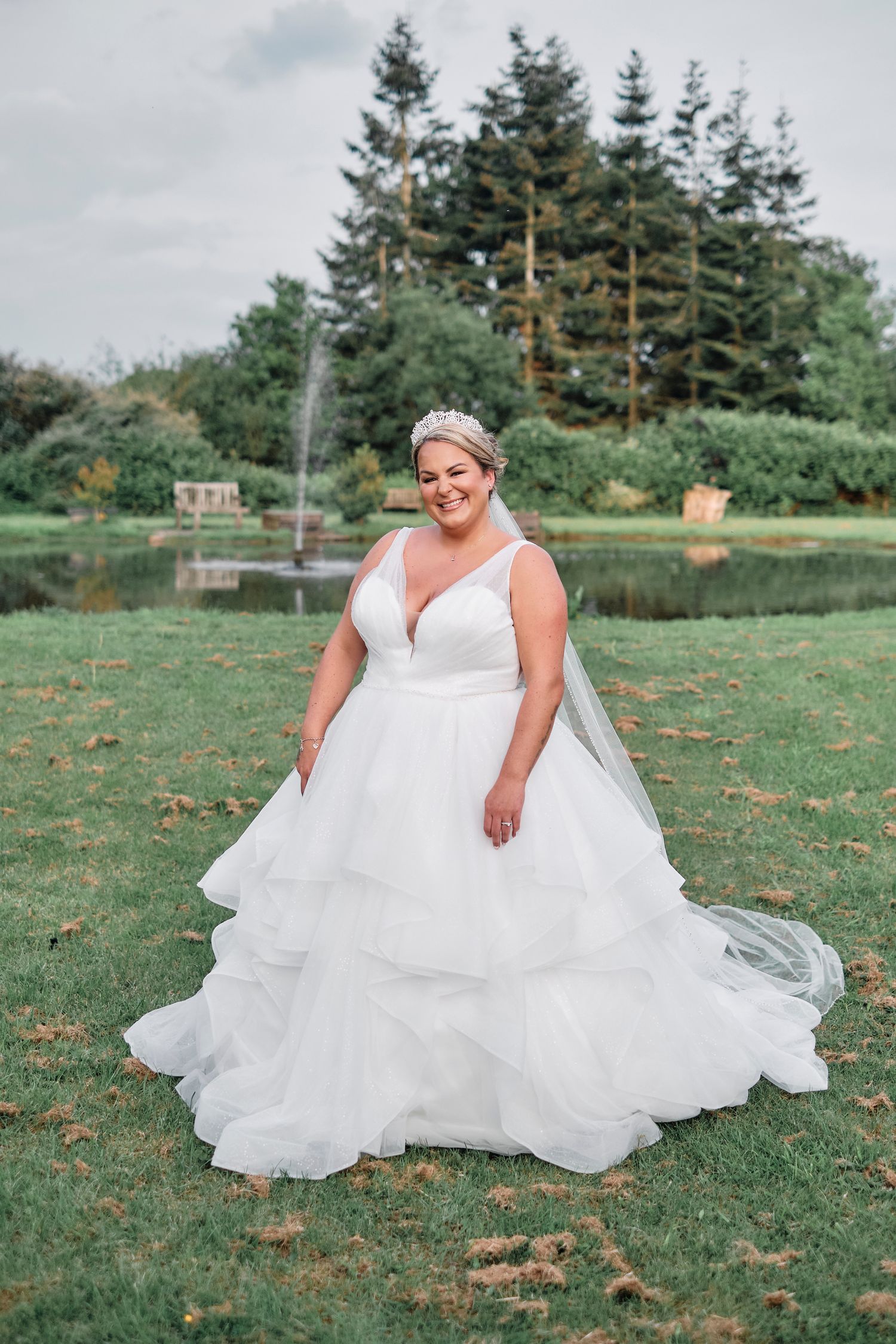 Plus-size bride smiles in layered tulle ballgown with tiara and veil in front of garden pond.
