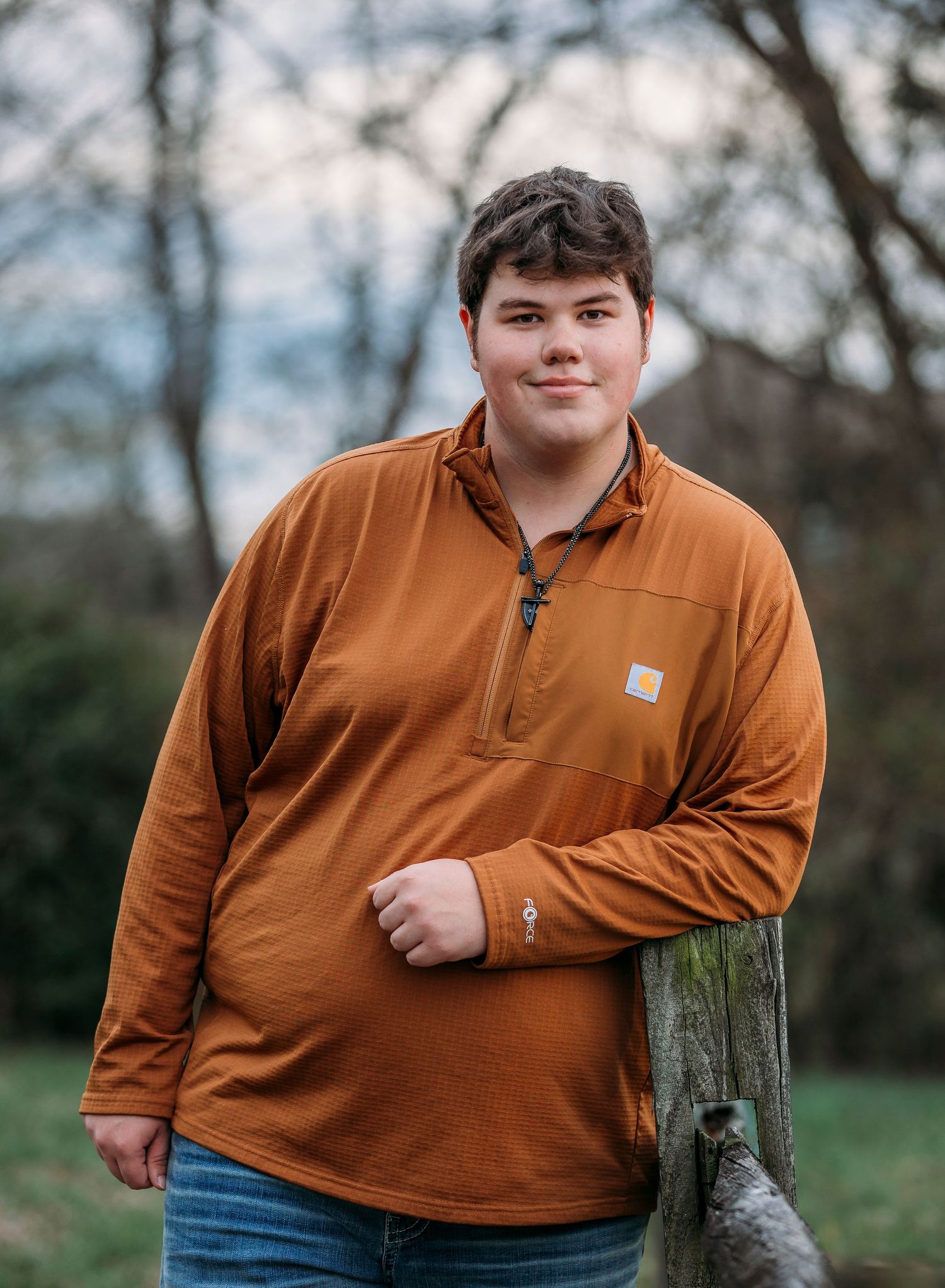 A person wearing an orange Carhartt quarter-zip pullover and jeans leans casually against a wooden post outdoors.
