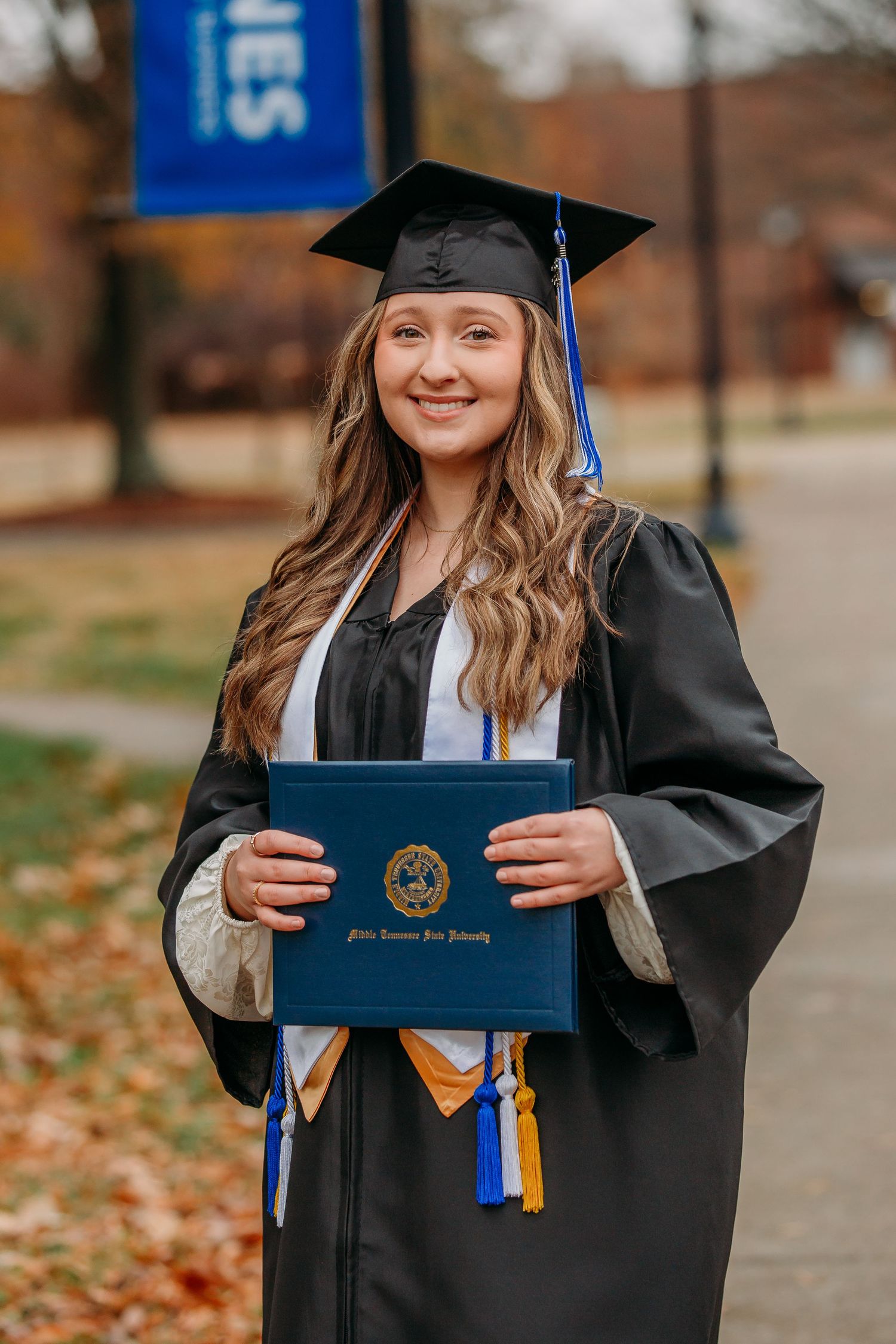 Graduate in cap and gown poses with diploma on campus pathway lined with autumn leaves.