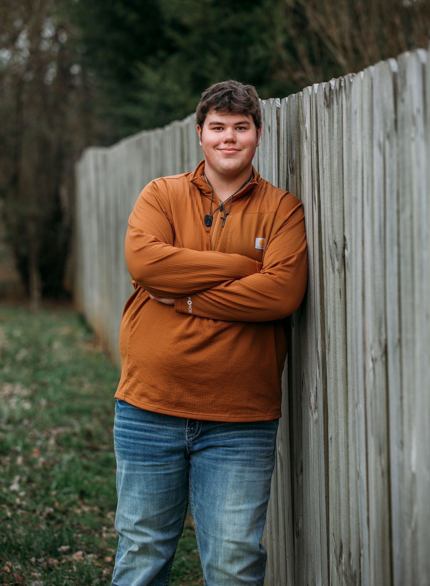 Person in brown jacket and jeans poses with crossed arms against white wooden fence.