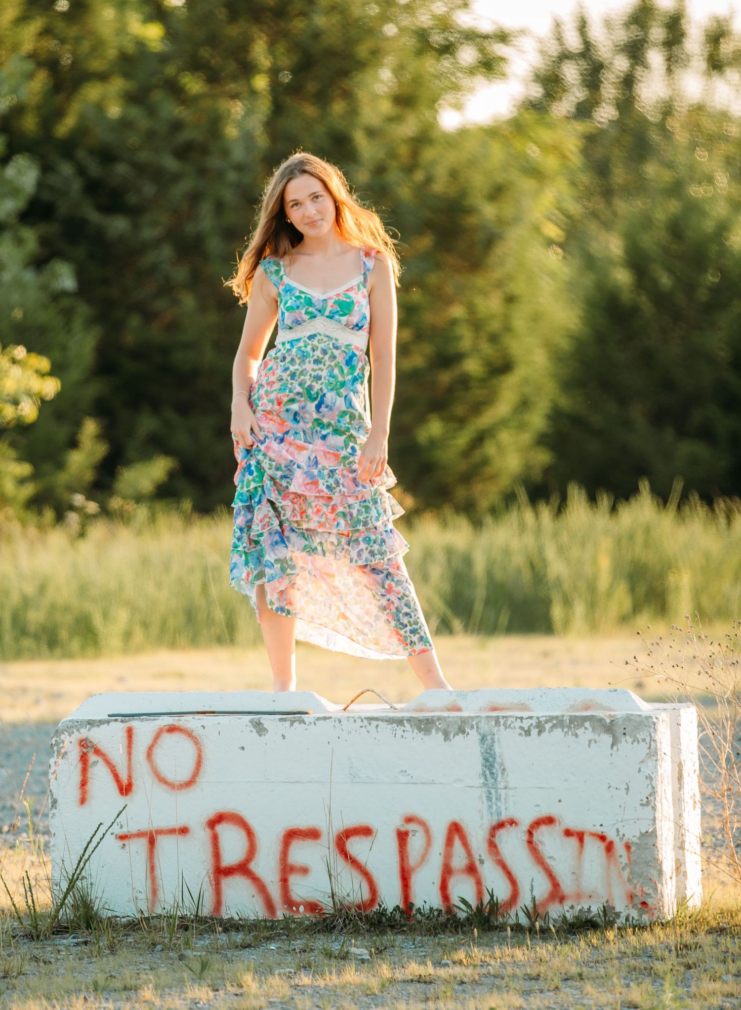 Person in floral dress stands on concrete barrier with No Trespassing graffiti at sunset.
