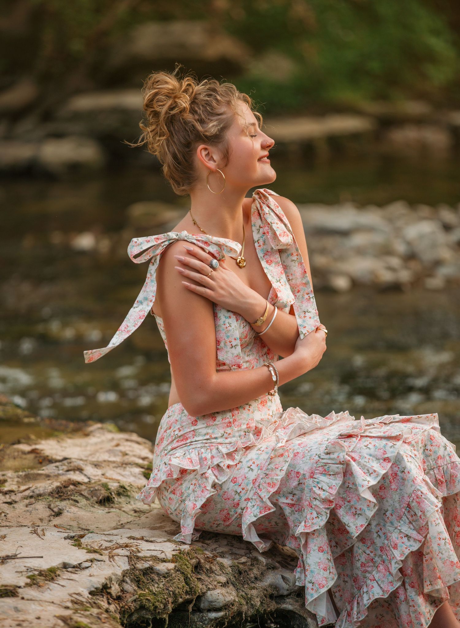 Person in a floral dress sitting on rocky riverbank during a summer photo shoot in nature.