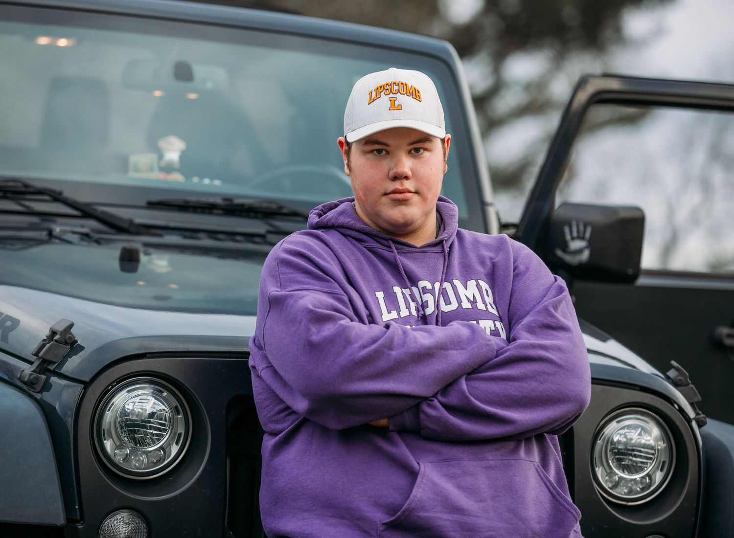 A person in a purple sweatshirt and baseball cap stands with crossed arms in front of a black Jeep.