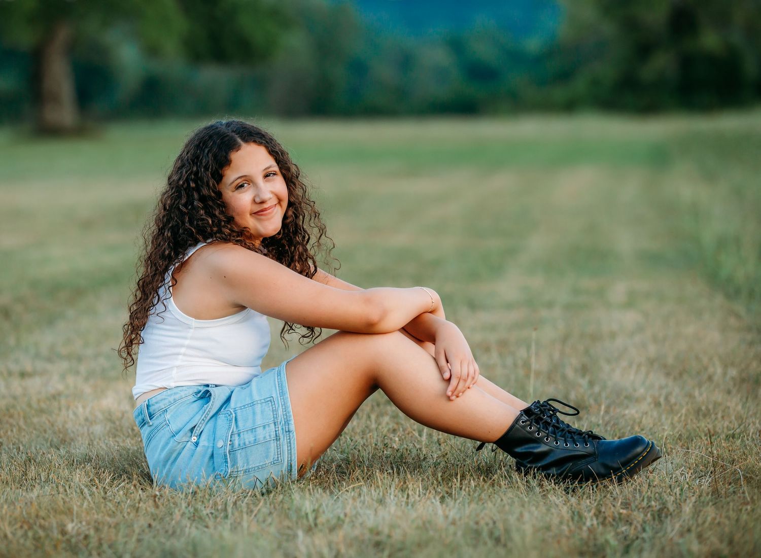 A person sits casually on grass wearing denim shorts, a white tank top, and black boots in a sunny outdoor setting.