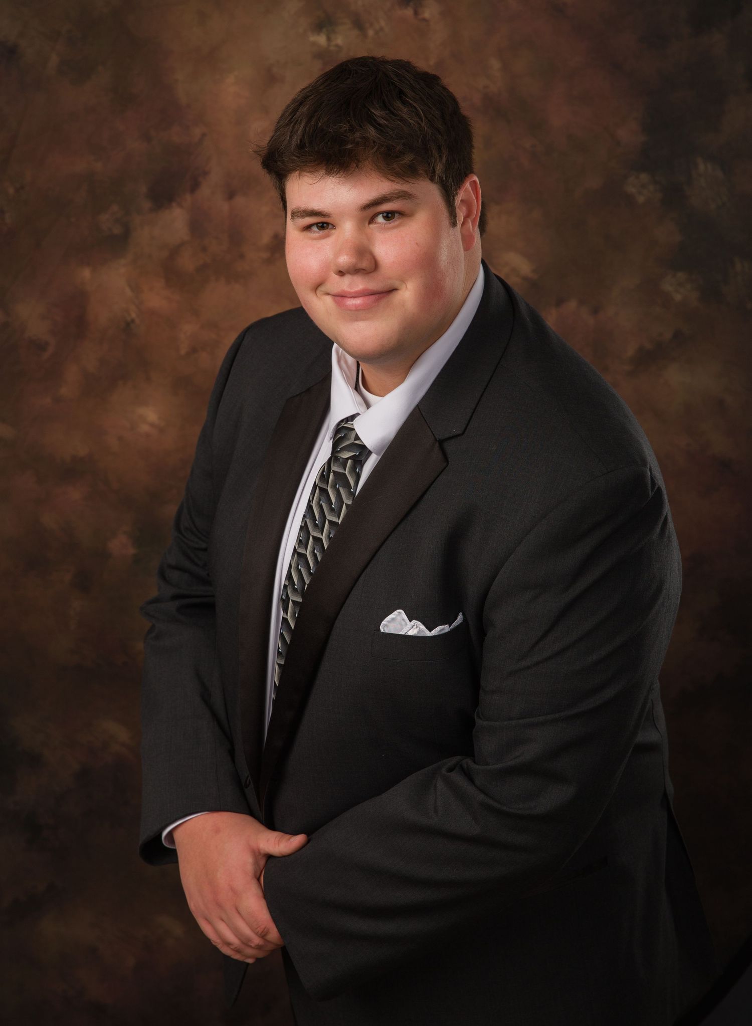 Professional studio portrait of someone in formal black suit and striped tie against brown mottled backdrop.