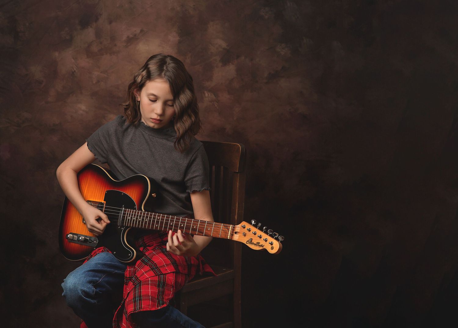 A musician in a gray t-shirt plays an acoustic sunburst guitar against a dark brown backdrop.