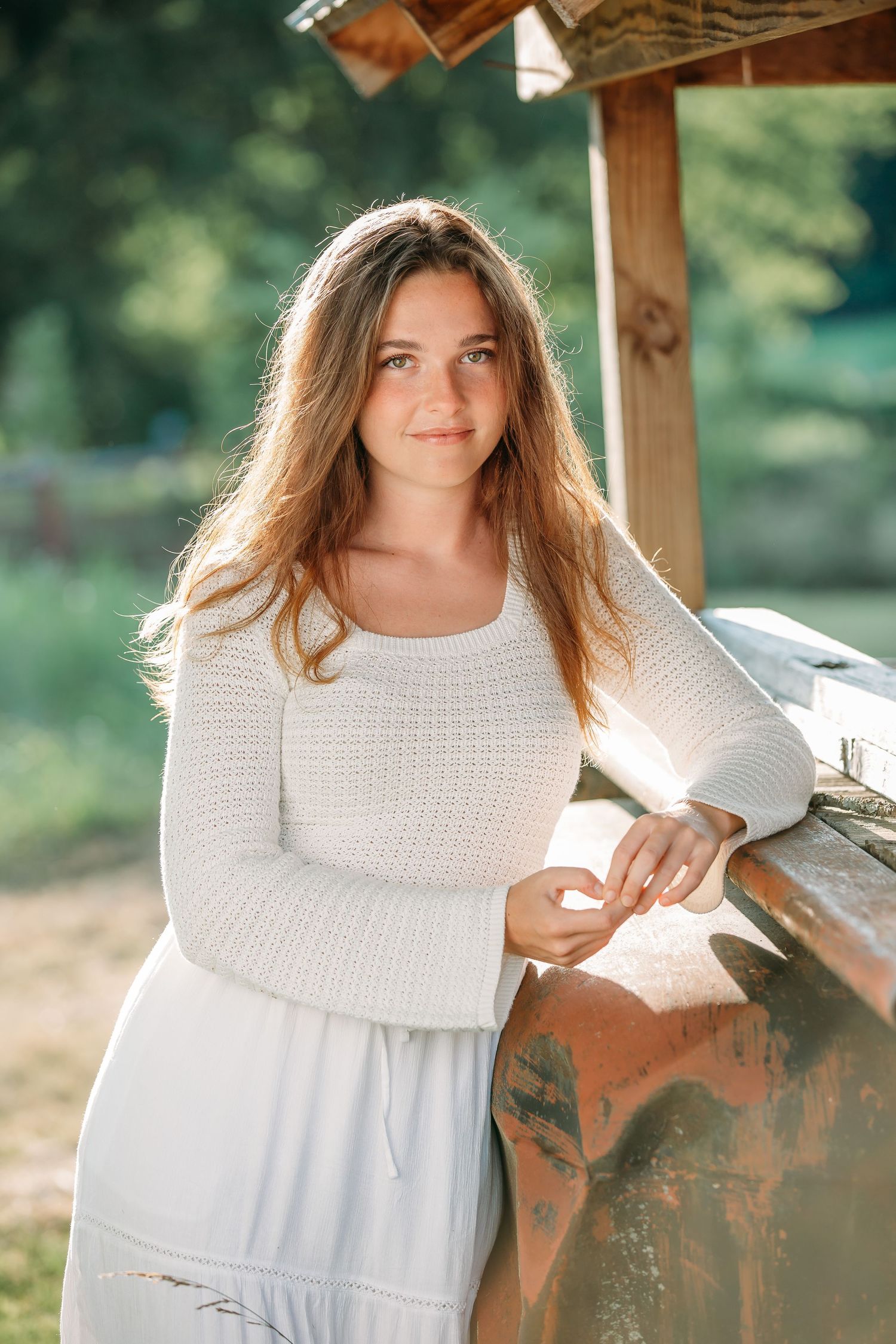 A person in a white knit sweater and flowing dress poses by a rustic wooden structure in natural lighting.