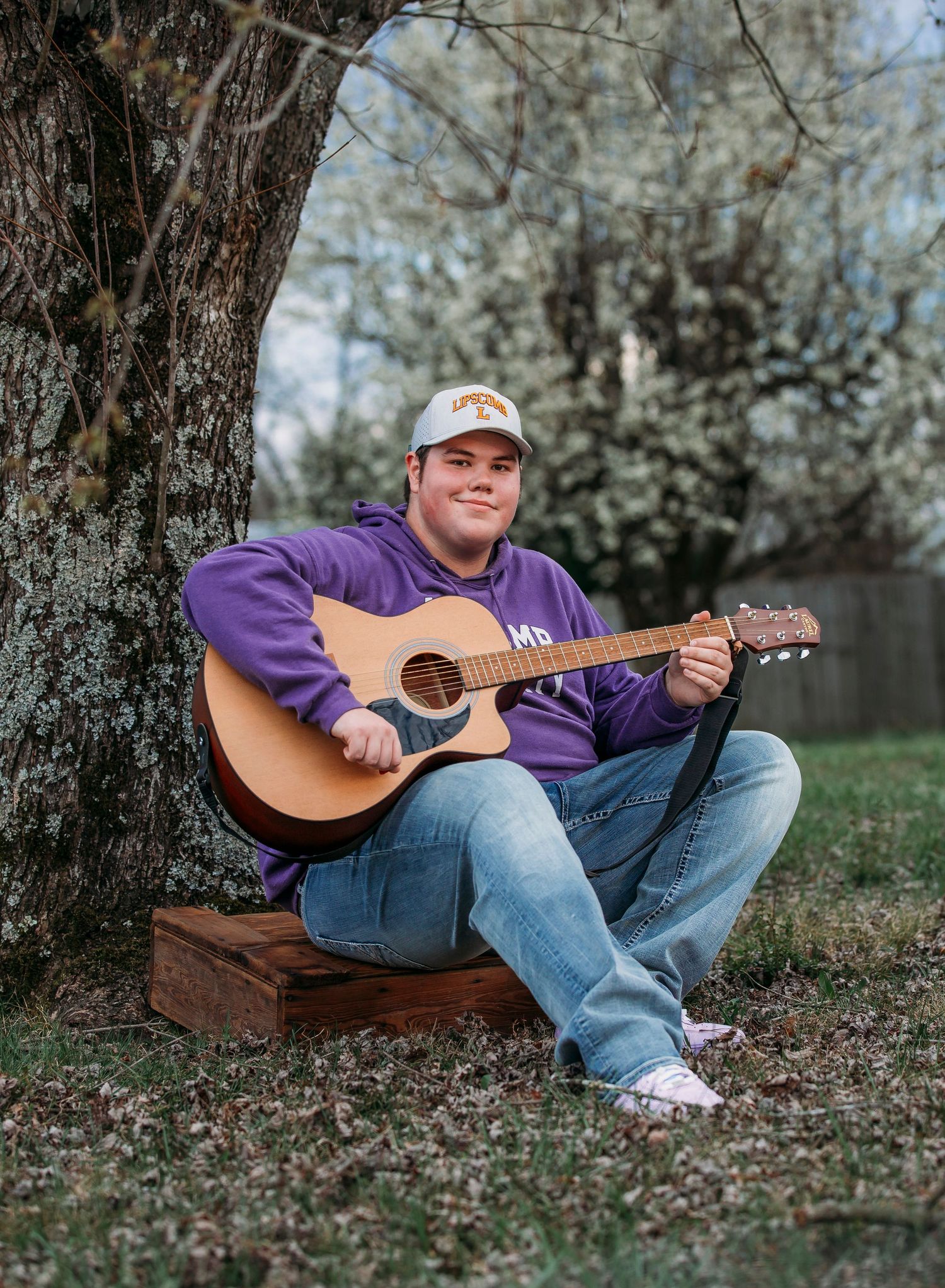 A person in jeans and purple shirt sits on grass playing an acoustic guitar among spring blossom trees.