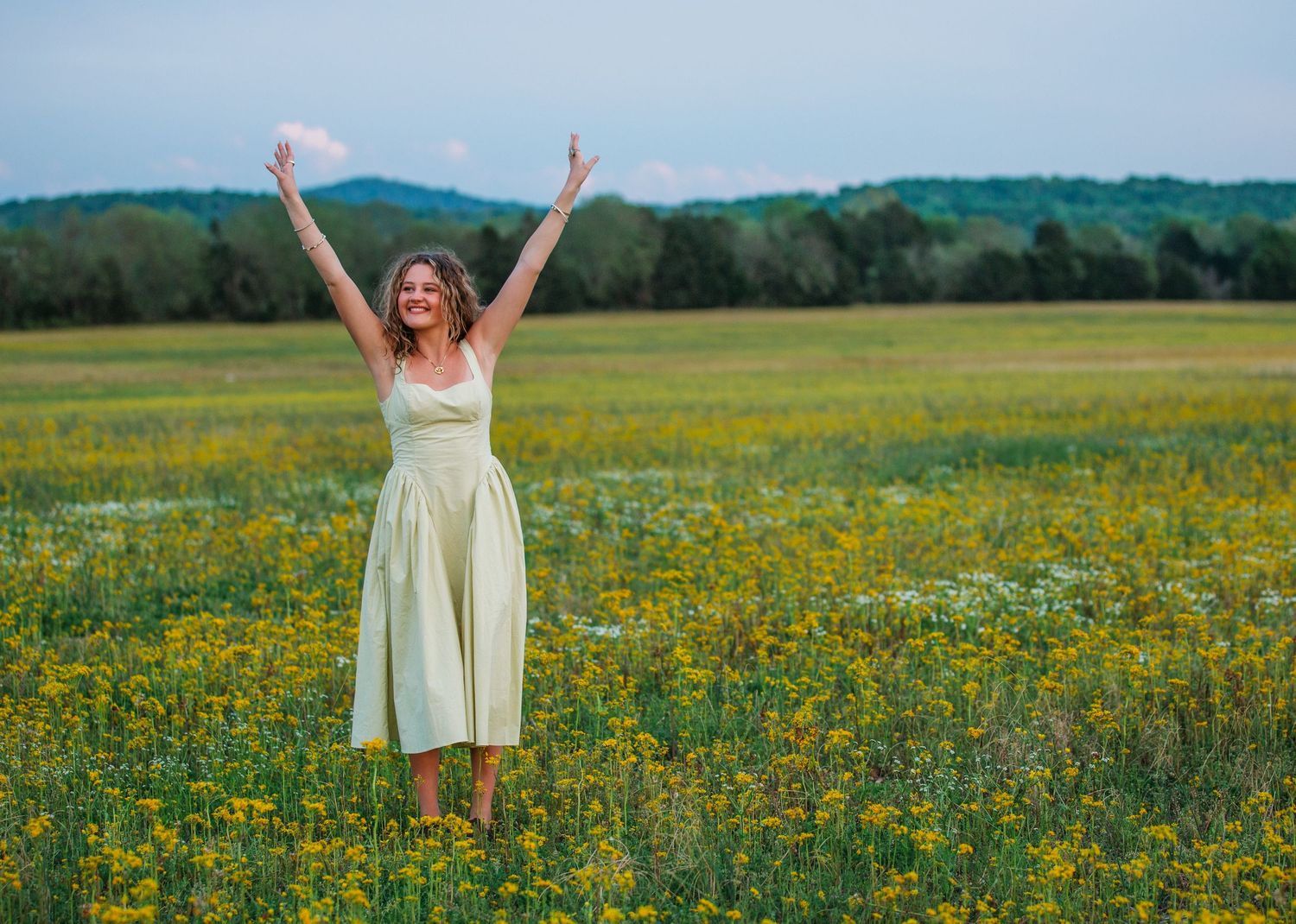 Person in yellow sundress stands joyfully with raised arms in a blooming wildflower meadow against green rolling hills.