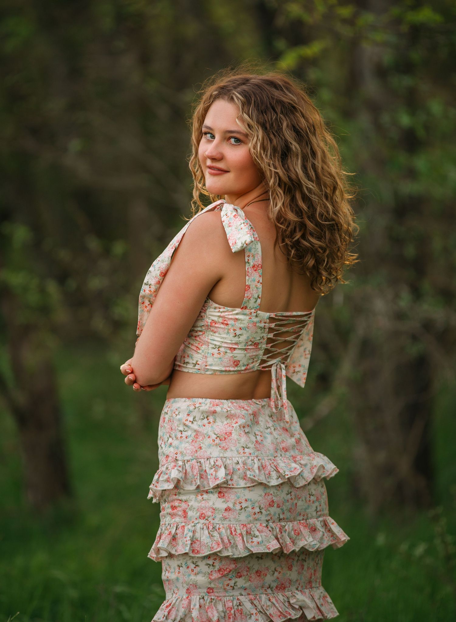 A person in a floral ruffled sundress twirls playfully in a lush green outdoor setting during summer.