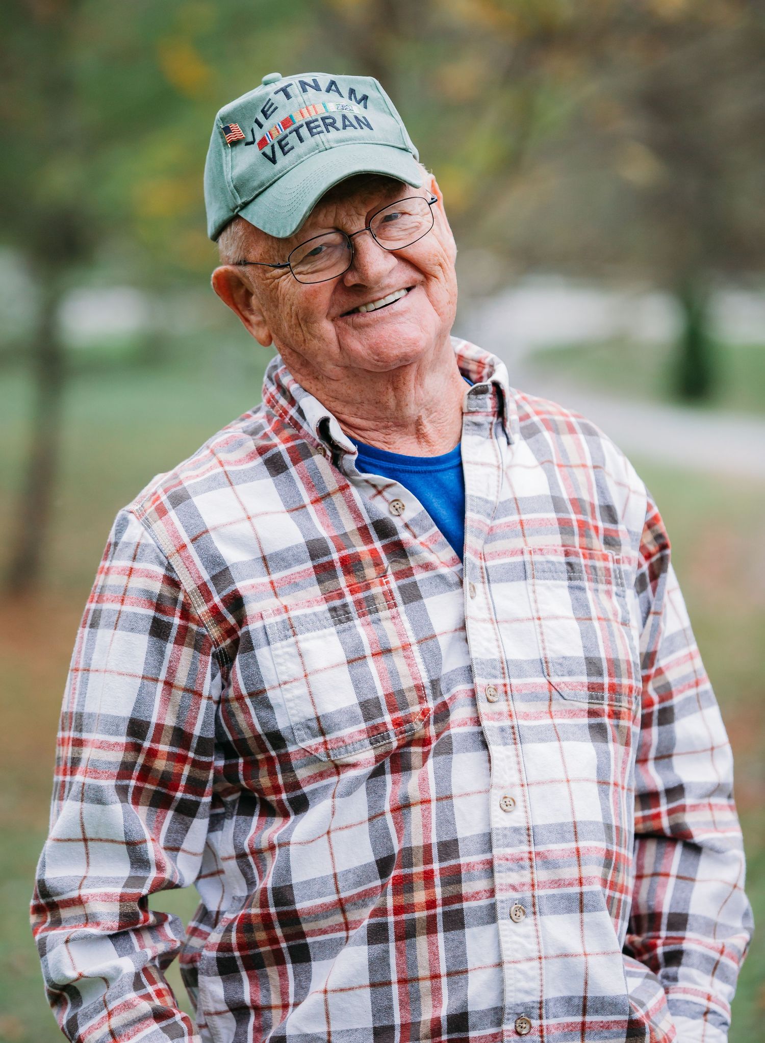 An elderly veteran wearing a Vietnam cap and plaid shirt smiles warmly outdoors.
