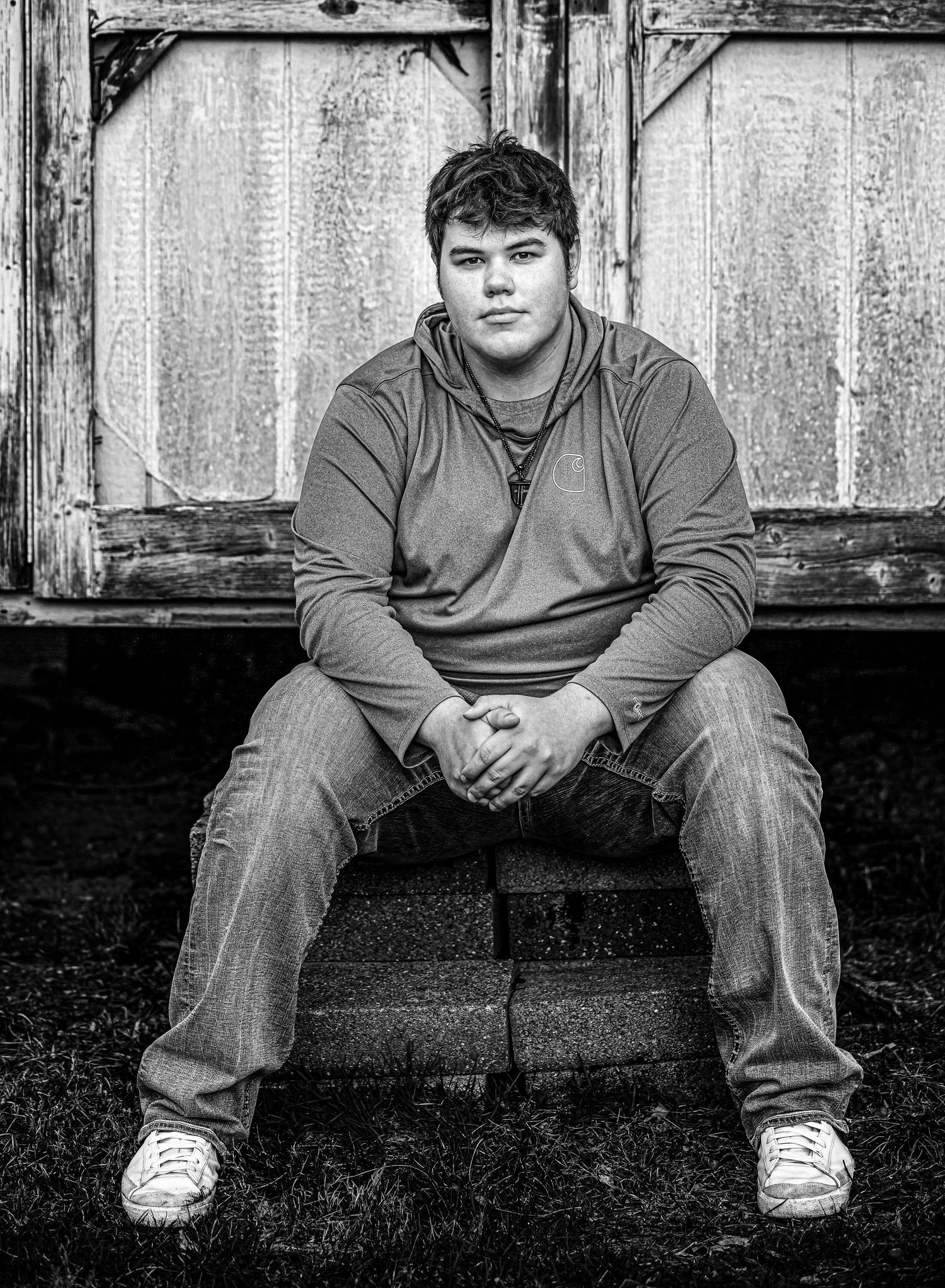 Black and white portrait of young man sitting on steps against weathered wooden wall.