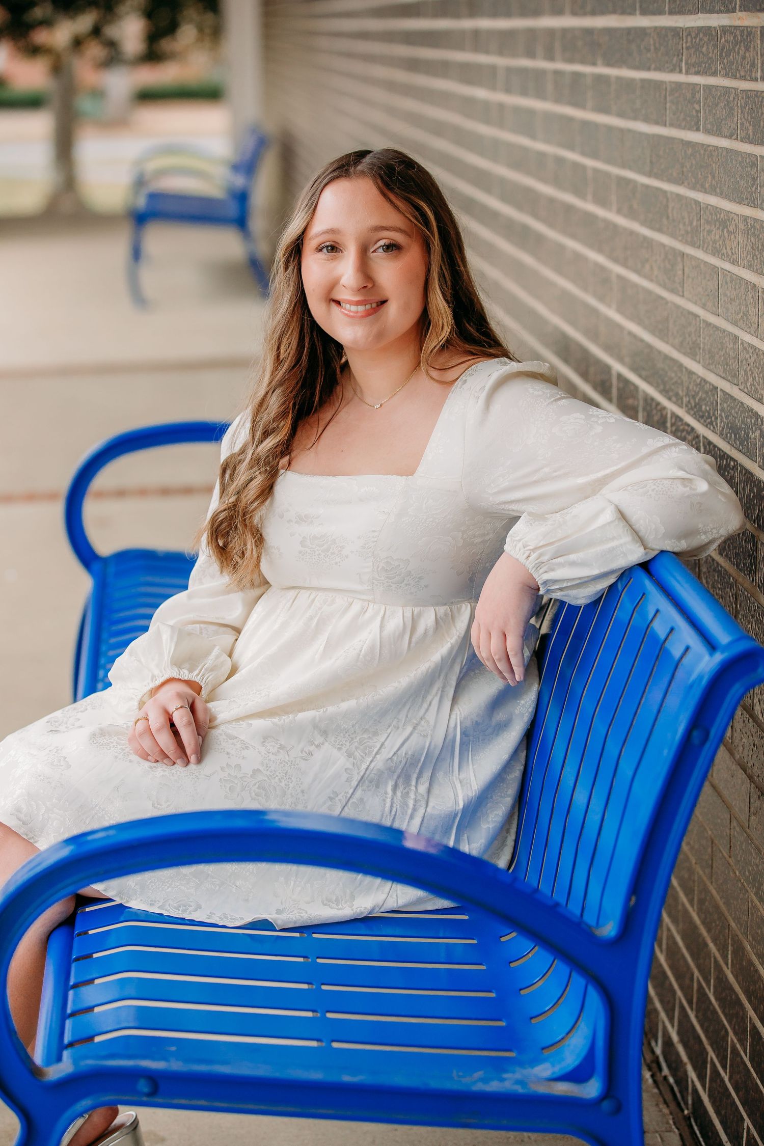 Person in a white dress sitting on a bright blue bench against a brick wall outdoors.