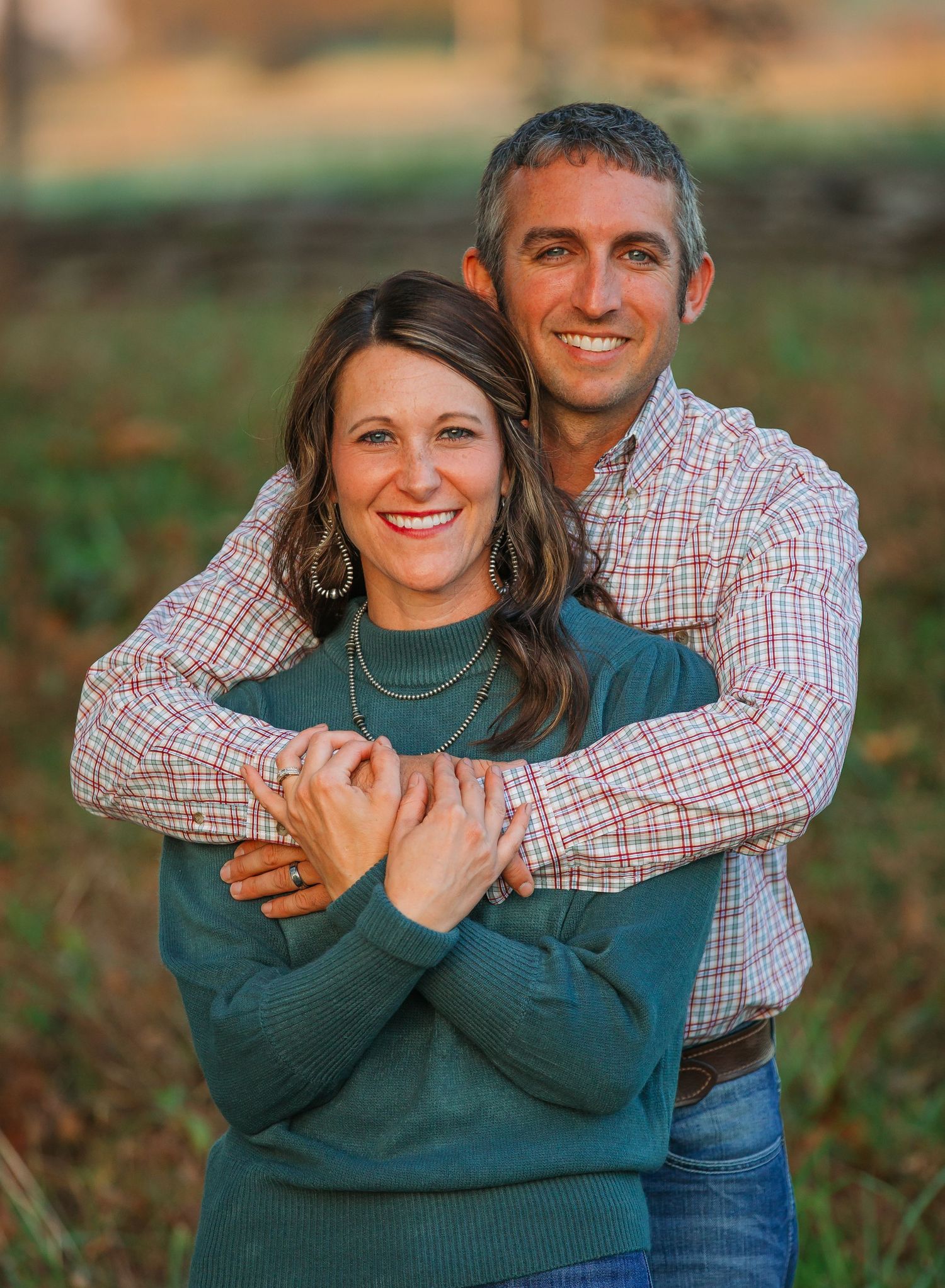 A couple embraces while wearing matching plaid shirts and standing in an autumn field.