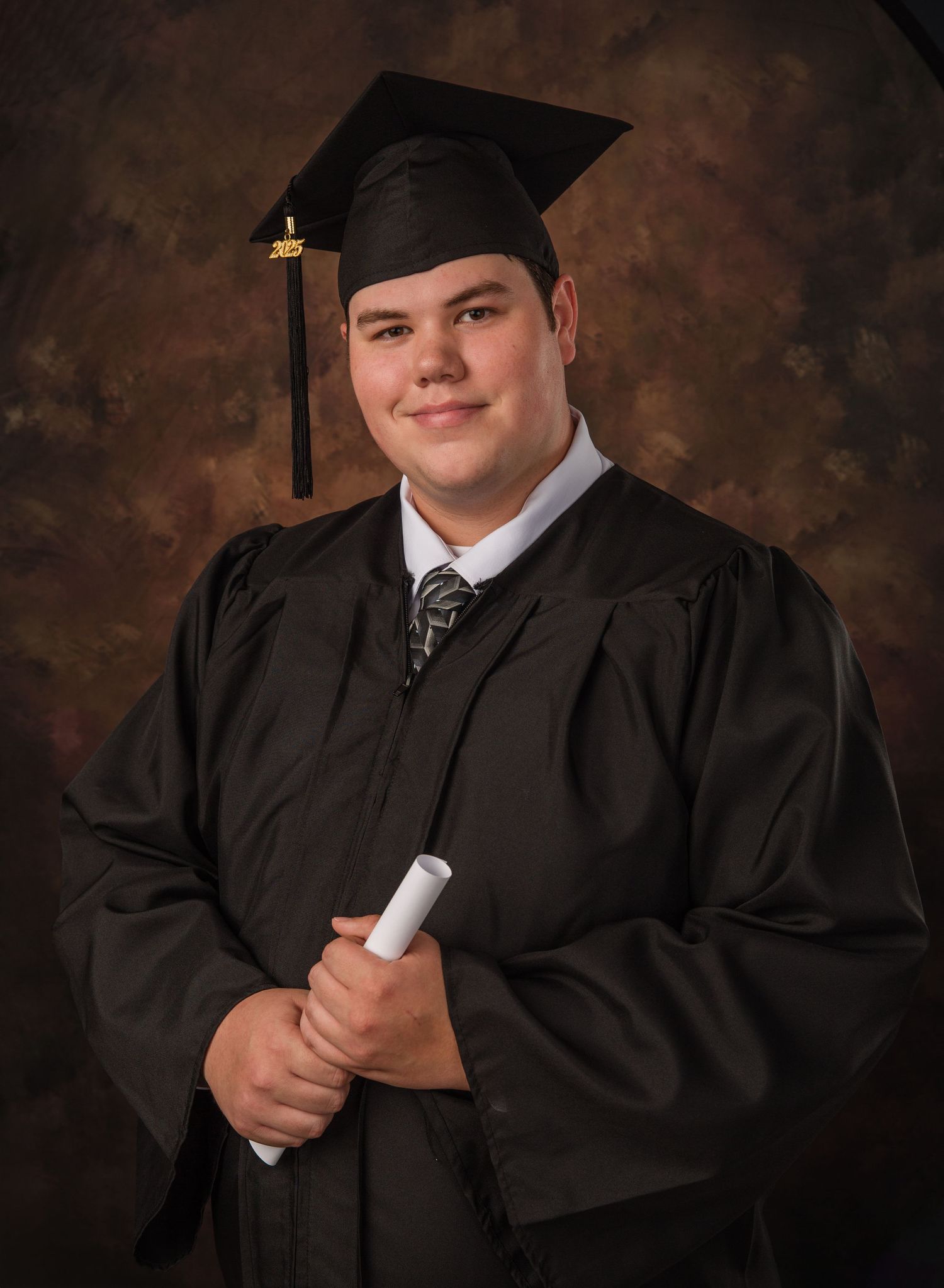 A graduate in cap and gown poses with diploma during formal portrait session against dark background.