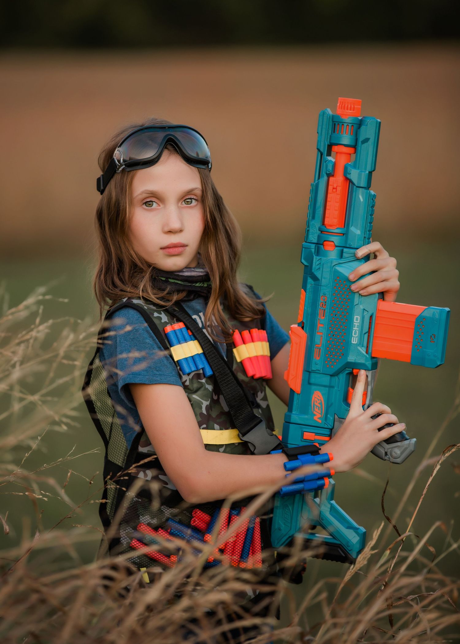 A person in tactical gear poses with a blue and orange toy gun in a field.