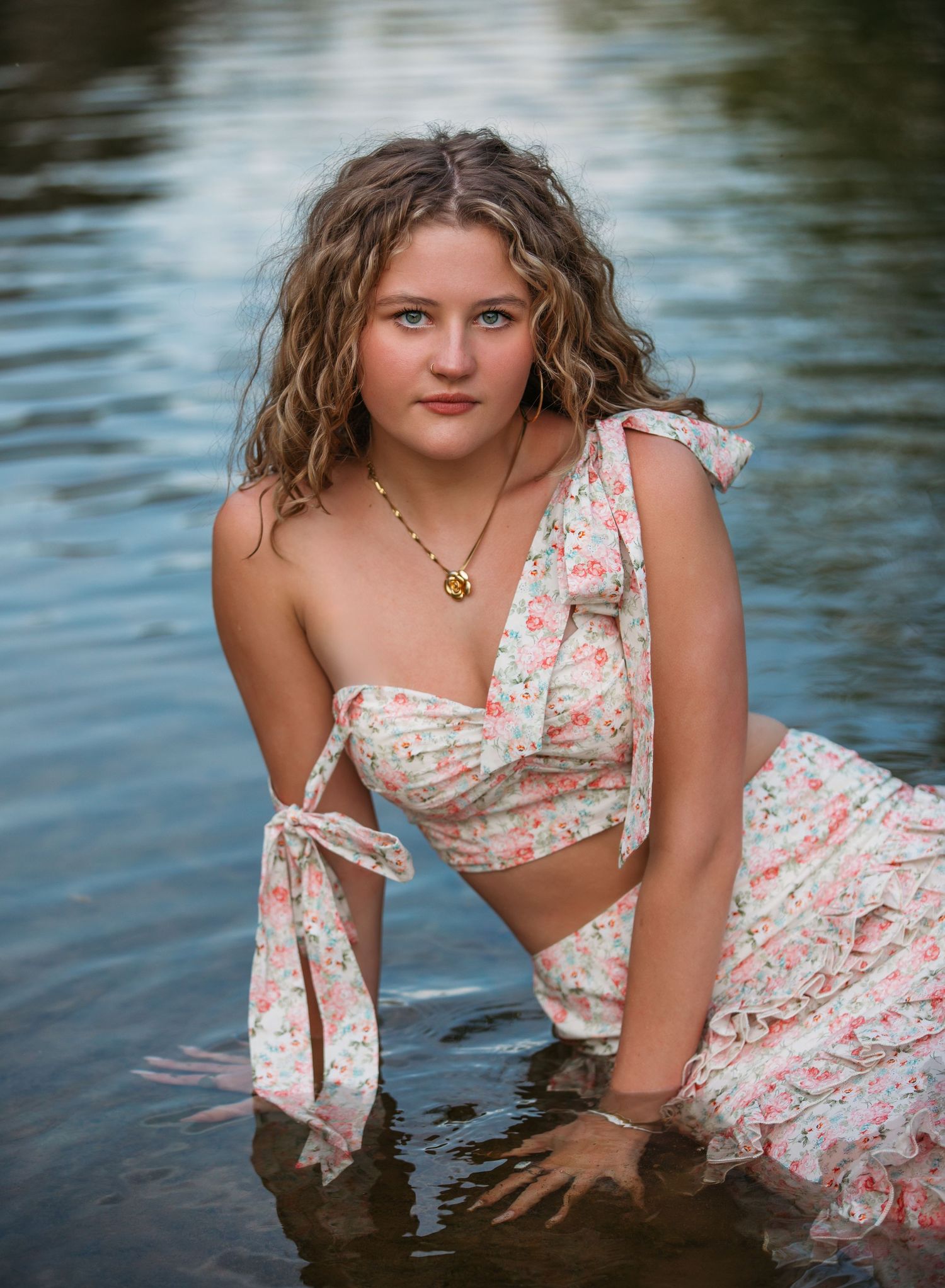 A woman in a floral dress crouches in calm water during a summer photoshoot.