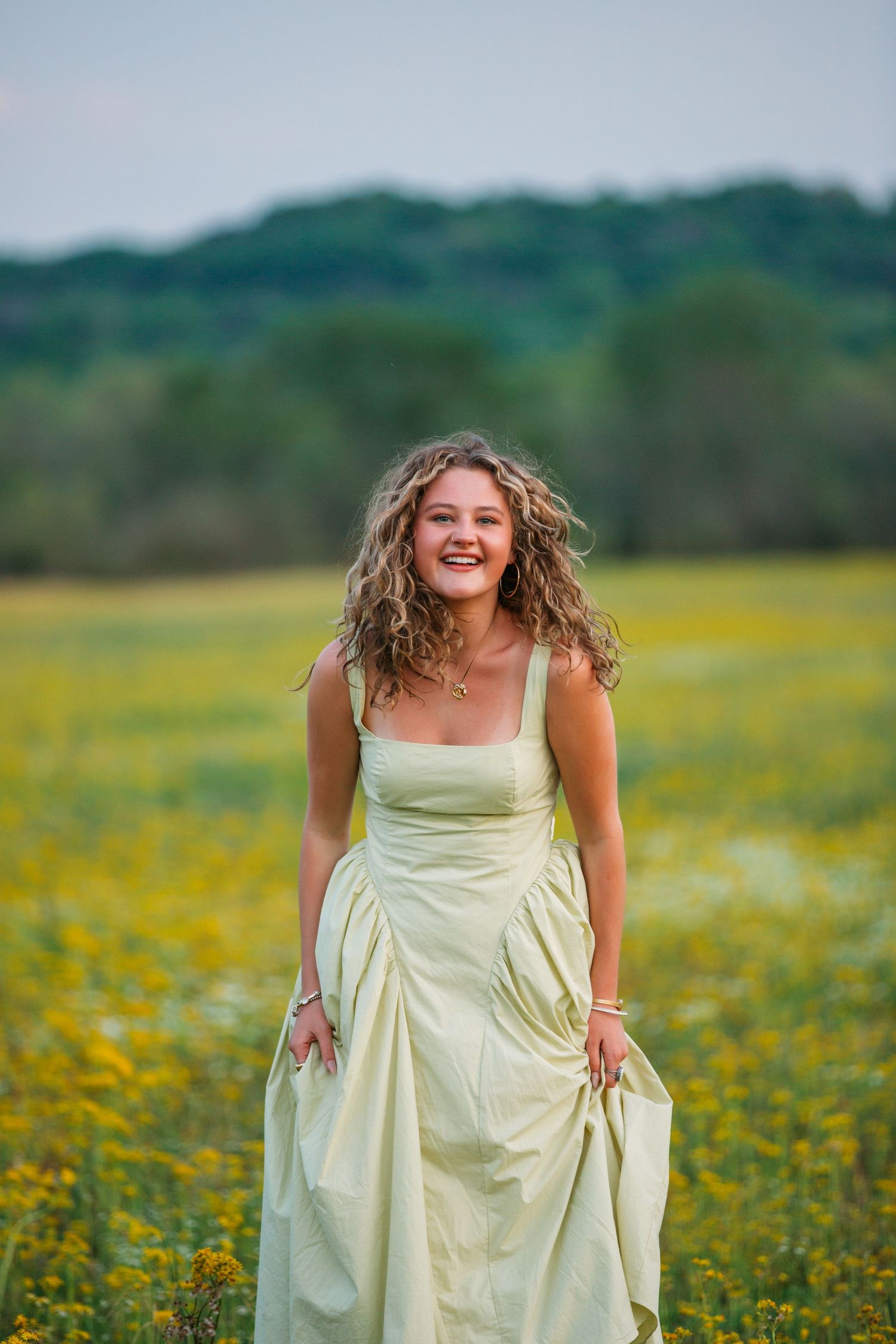 Person in a light-colored dress laughs joyfully while standing in a blooming yellow wildflower meadow on a summer day.