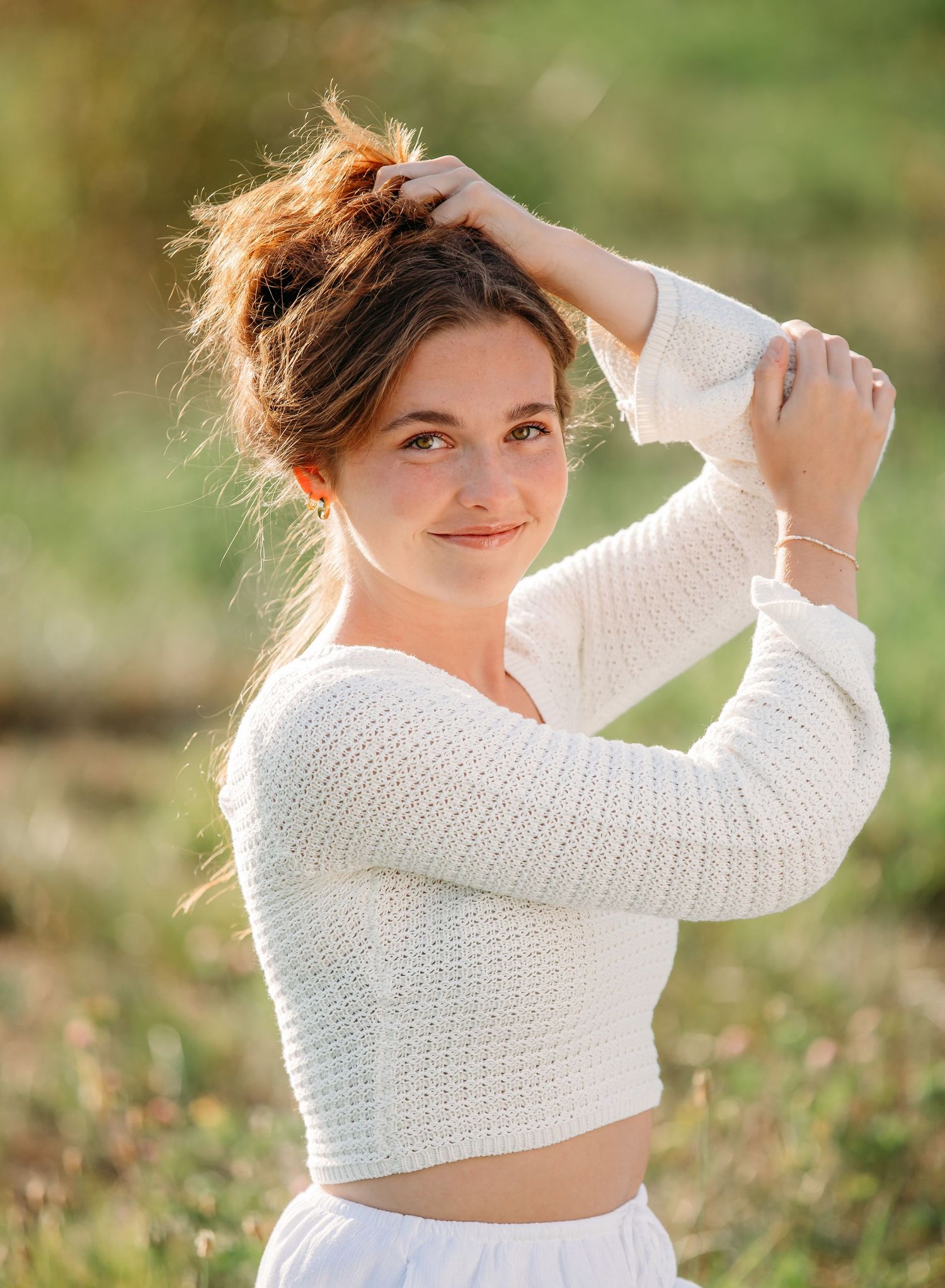 Someone wearing a white knit crop top poses outdoors with hands styled in their messy updo hairstyle.