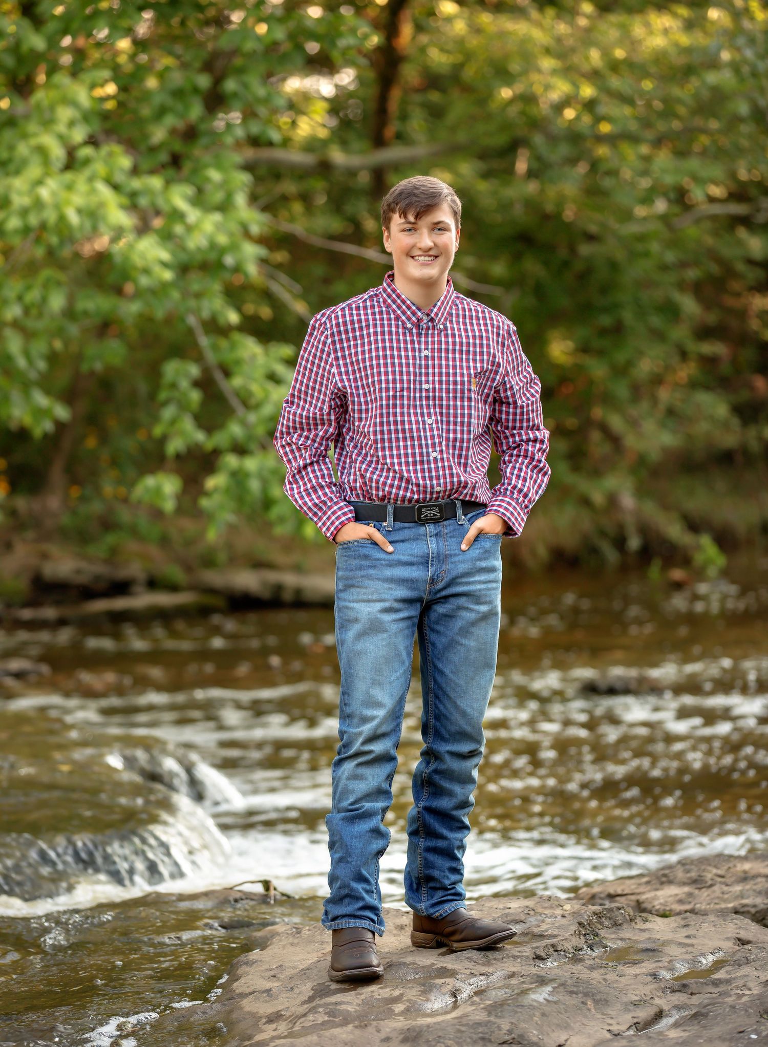 Person in red plaid shirt and jeans stands casually on rocky outcrop beside flowing stream in wooded setting.
