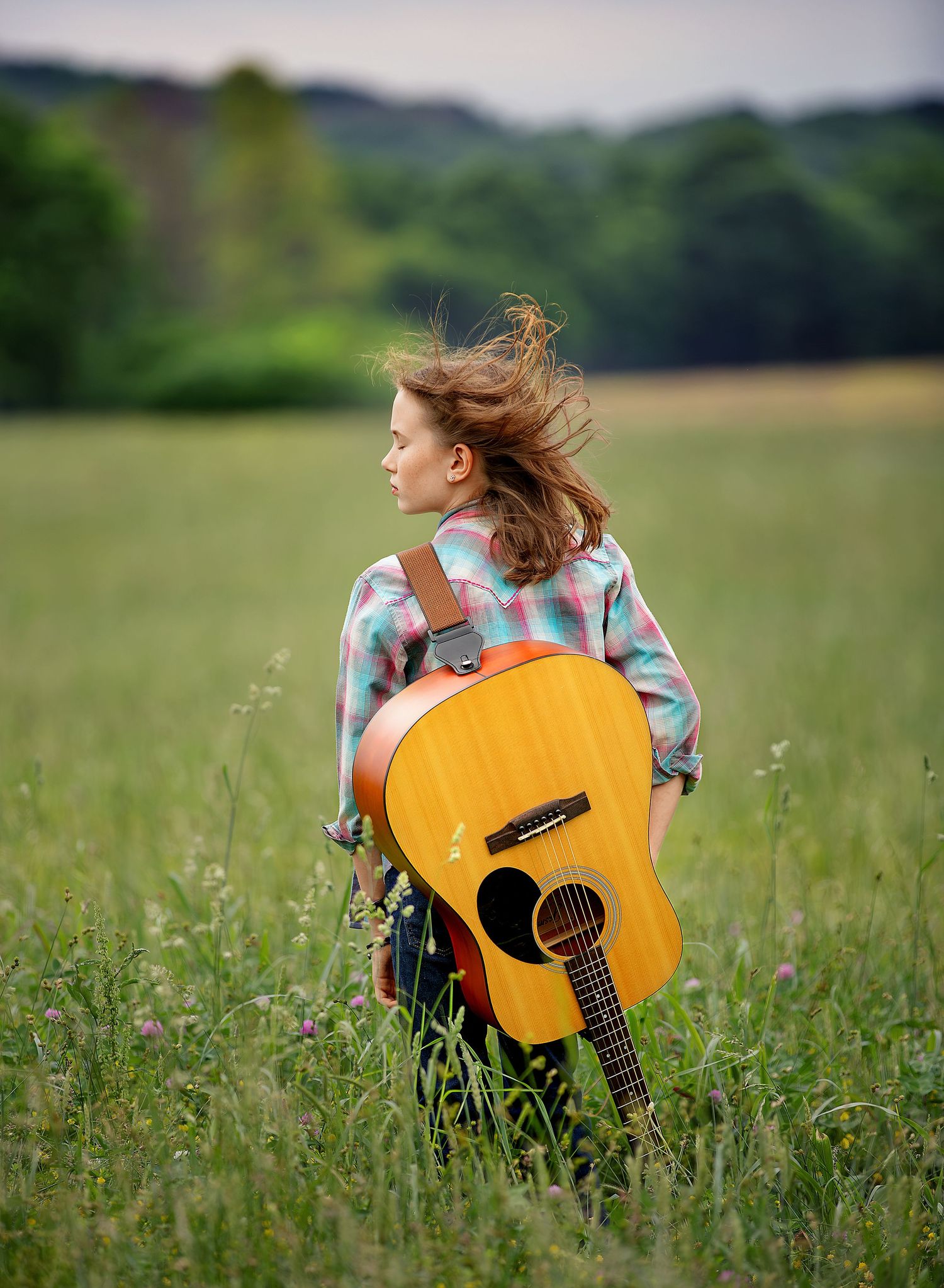 Someone sitting in a meadow holding a wooden acoustic guitar amidst tall grass and wildflowers on a sunny day.
