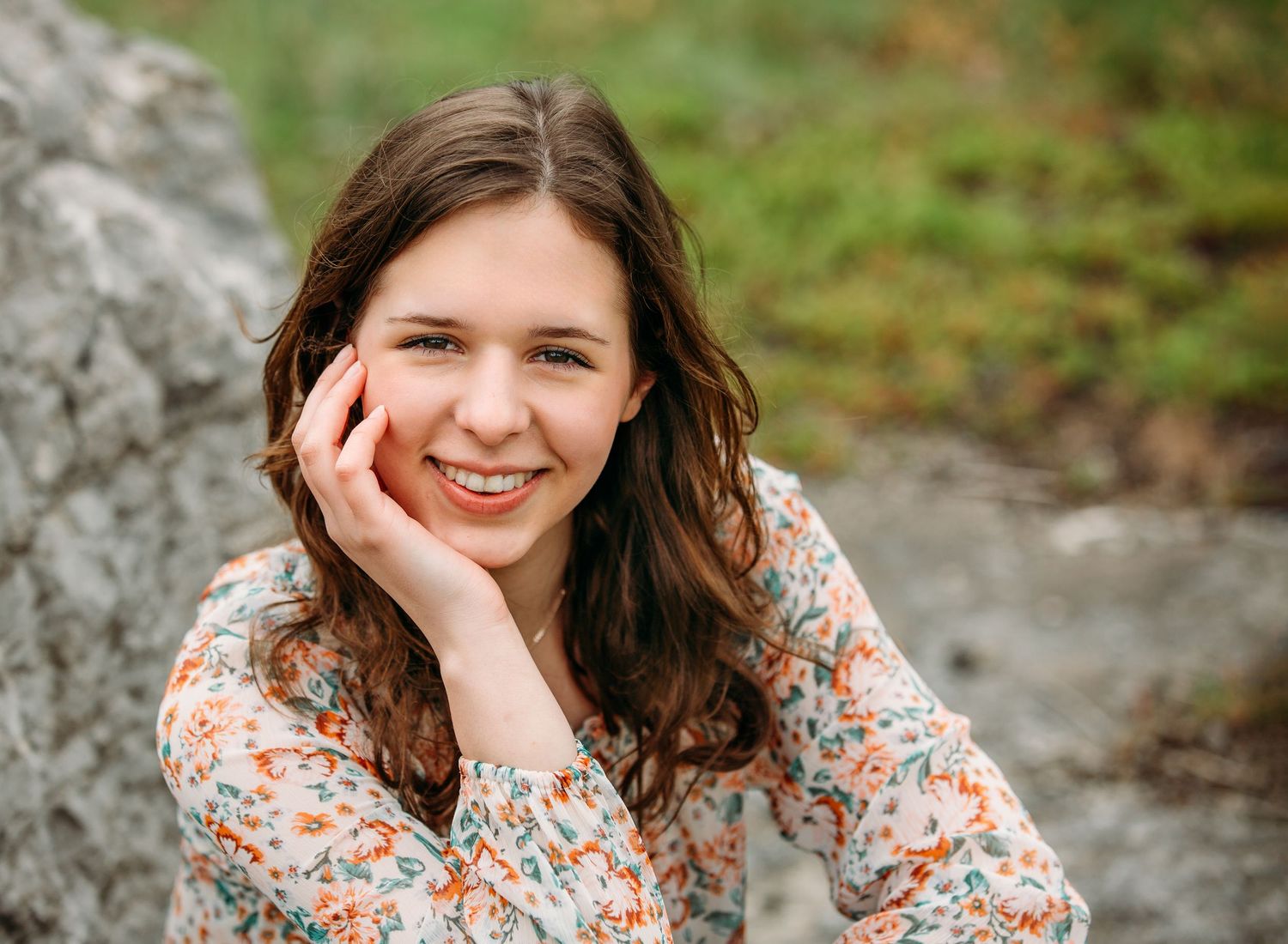 A person in a floral print top sits outdoors resting their chin on their hand against a natural rocky background.