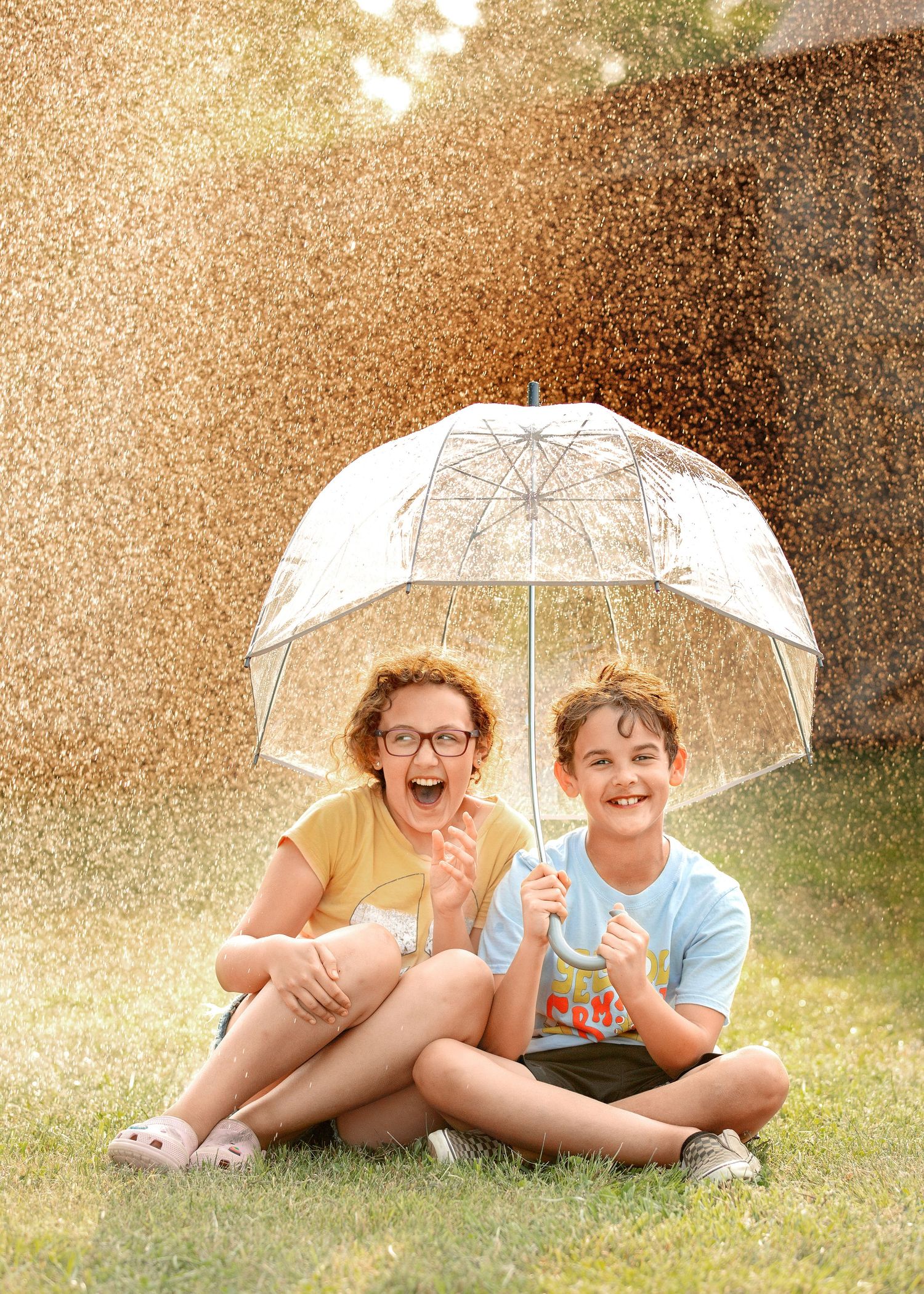 Two figures share a clear umbrella while sitting on grass during a sunlit rain shower.
