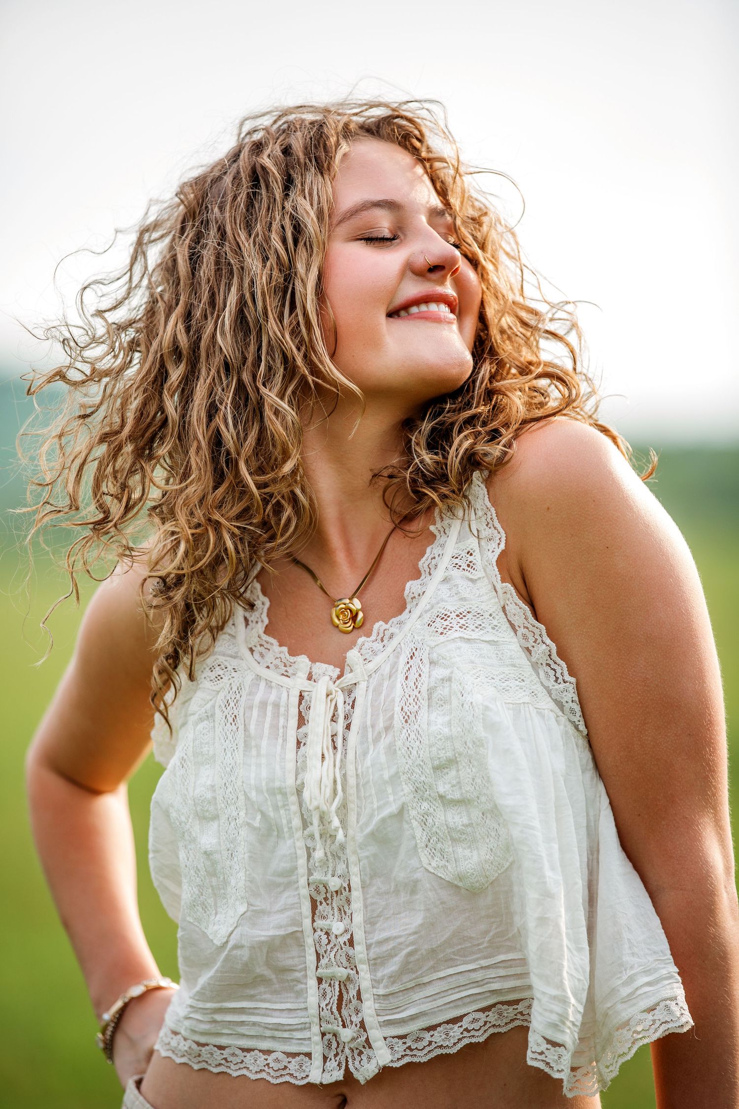 Woman with curly blonde hair laughs joyfully while wearing a white lace top outdoors on a sunny day.