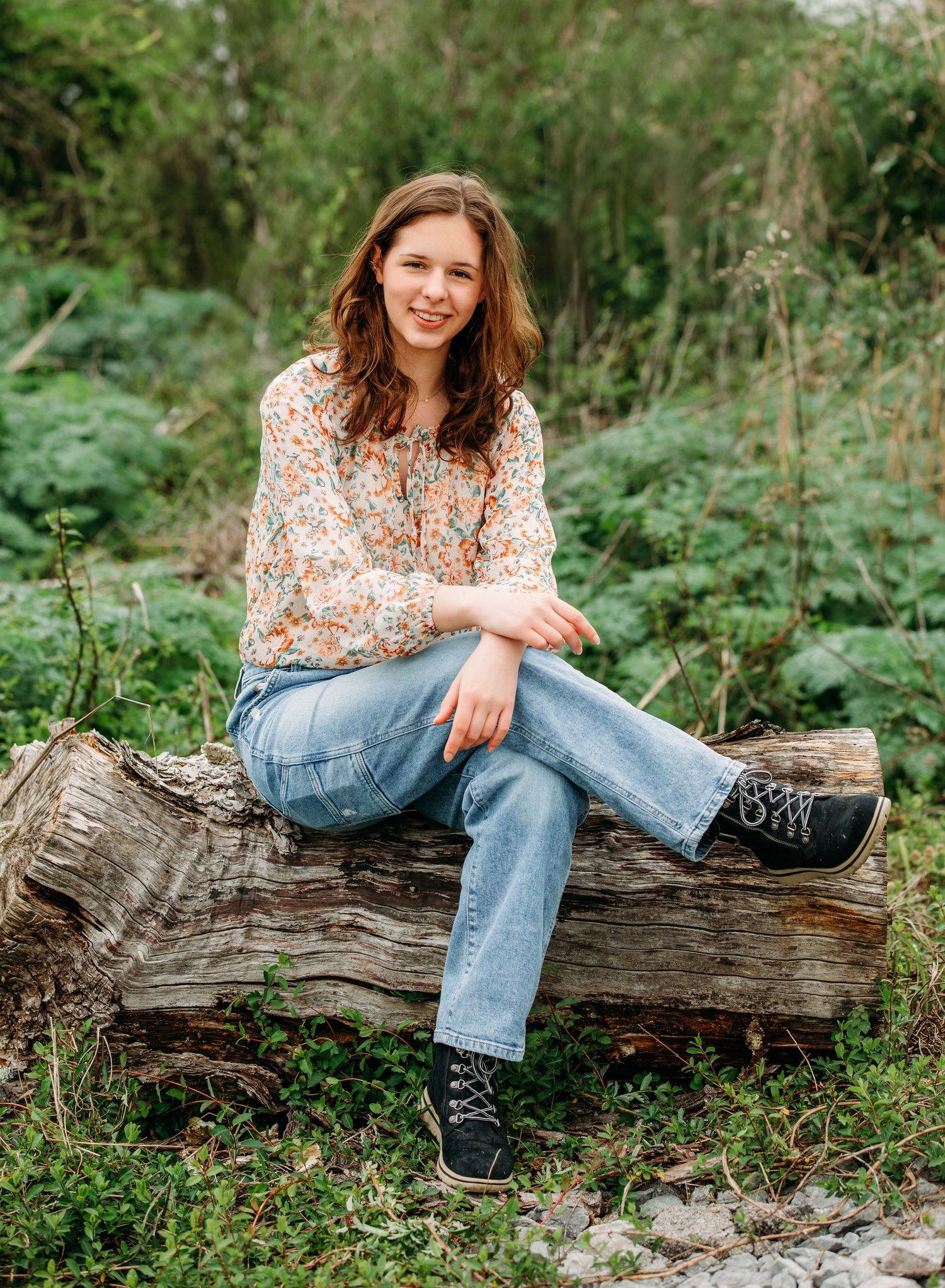 A person in a floral top and jeans sits casually on a fallen log surrounded by lush greenery in an outdoor setting.