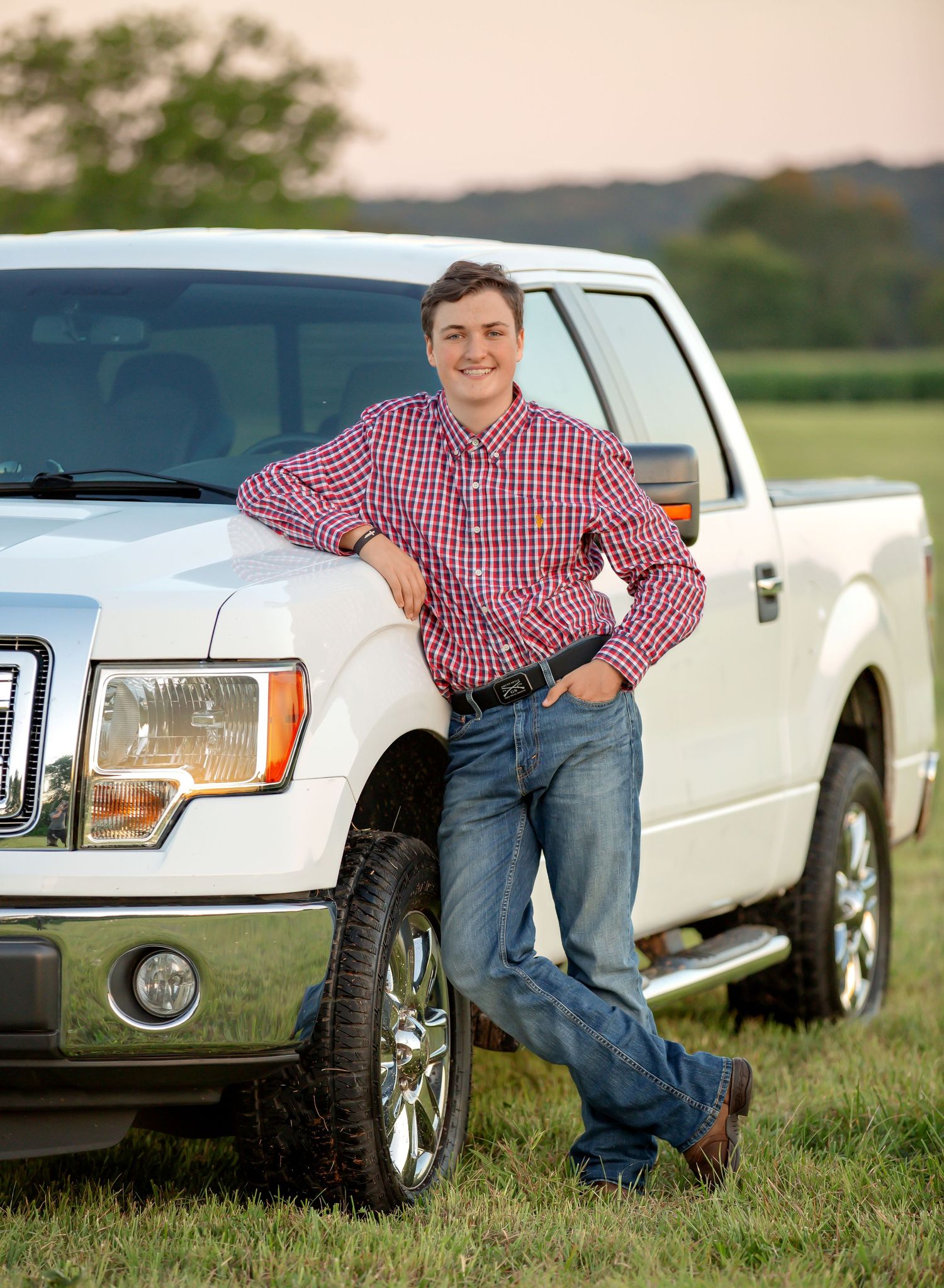 A person in a red plaid shirt leans casually against a white pickup truck parked on grassy terrain at sunset.