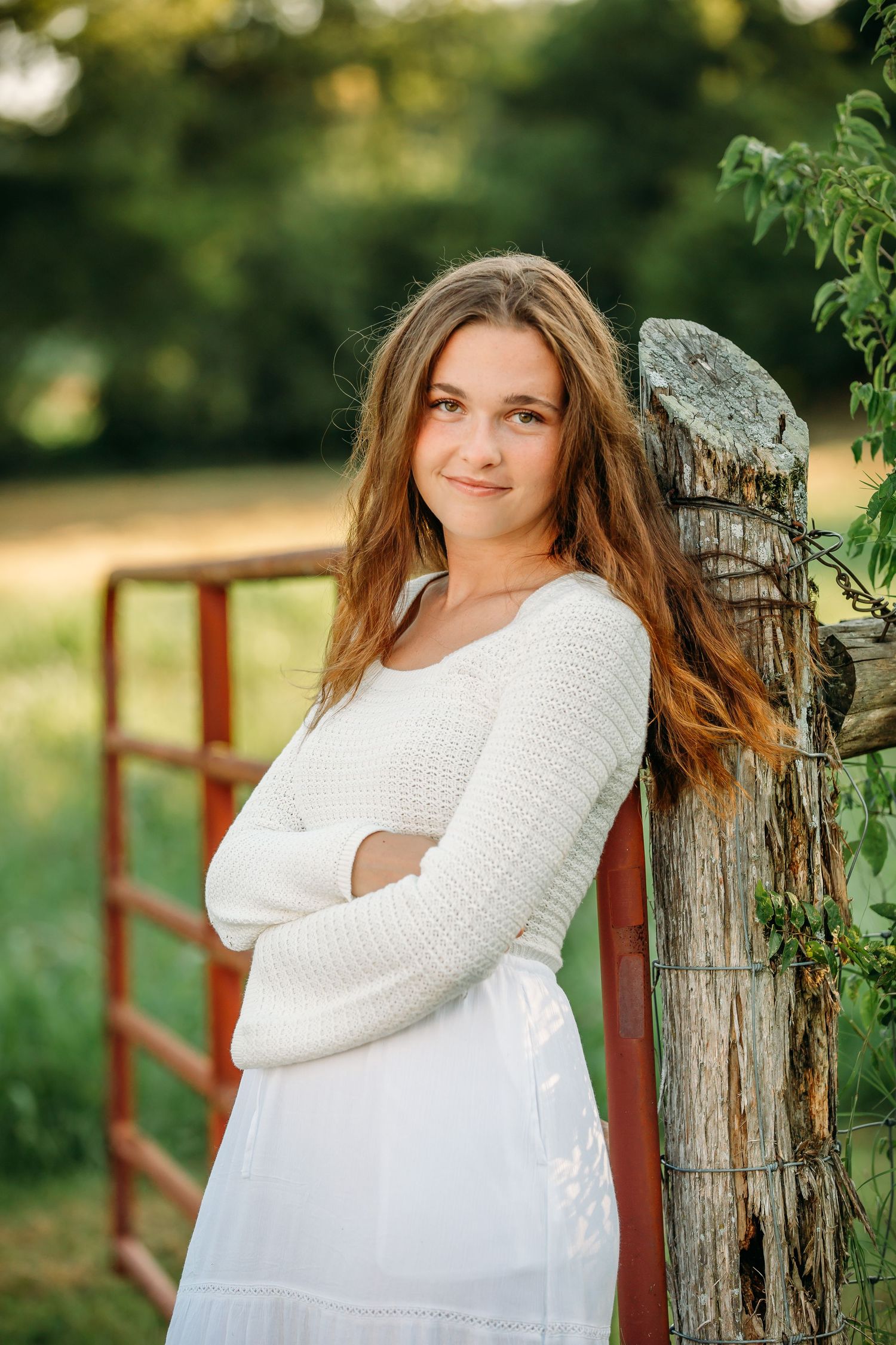 A person in a white sweater and dress leans against a rustic wooden fence post in a sunny outdoor setting.
