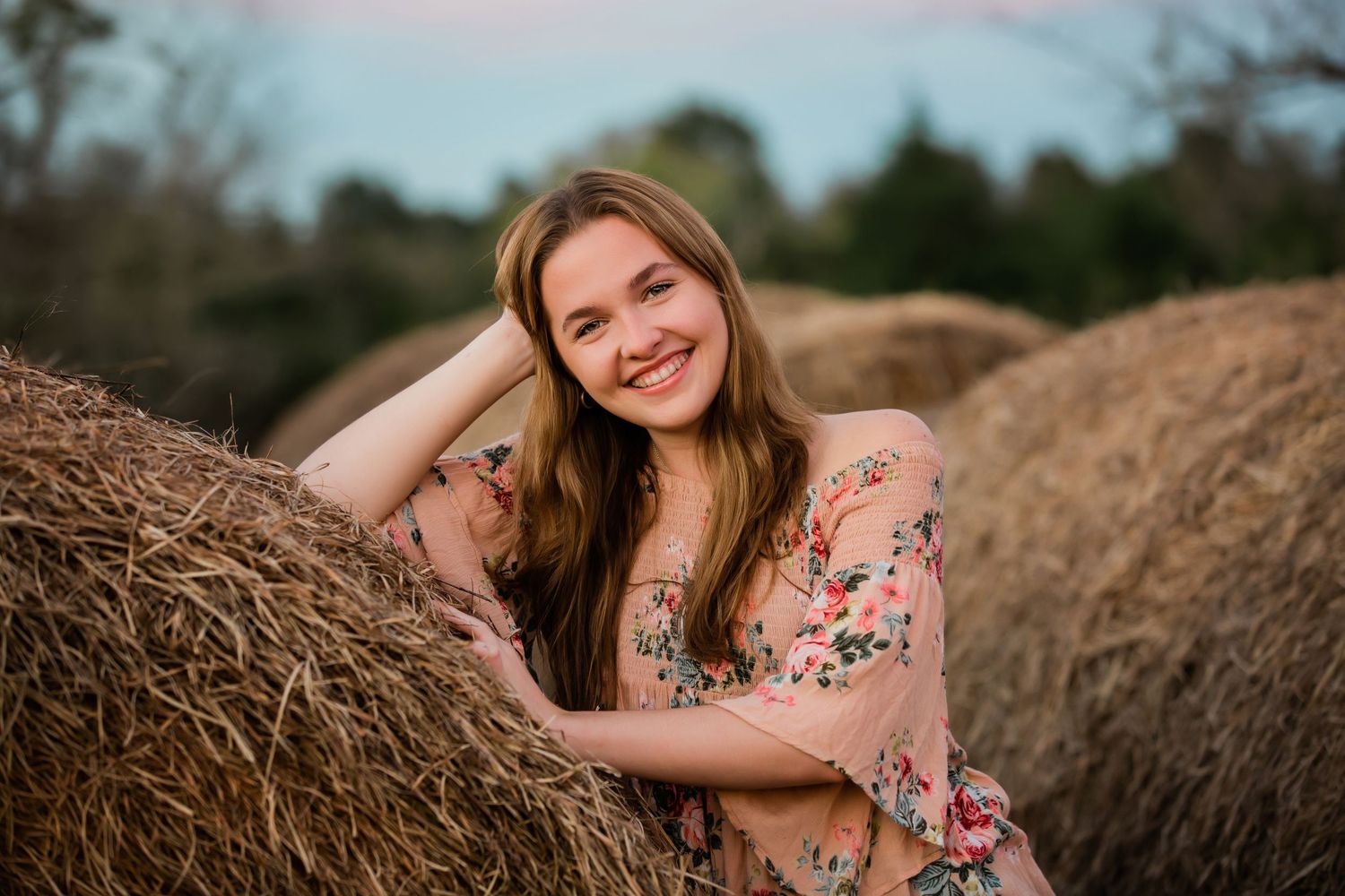 Portrait of someone in a floral dress posing against rustic hay bales during golden hour sunset.