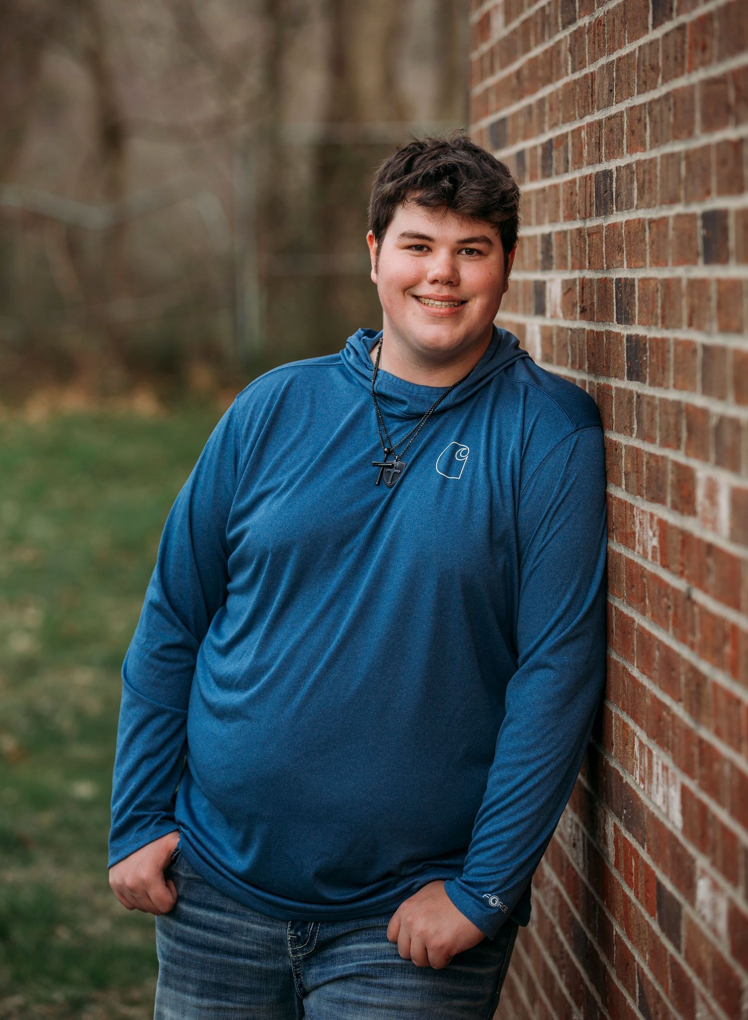 Student in blue long-sleeve shirt and jeans poses against brick wall in outdoor portrait series.