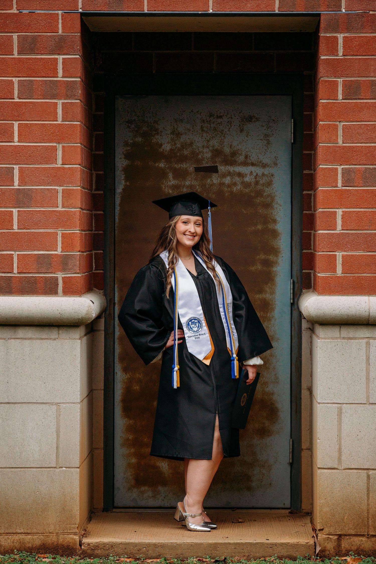Graduate in cap and gown poses in front of rustic metal door and brick wall wearing honors stole and graduation attire.