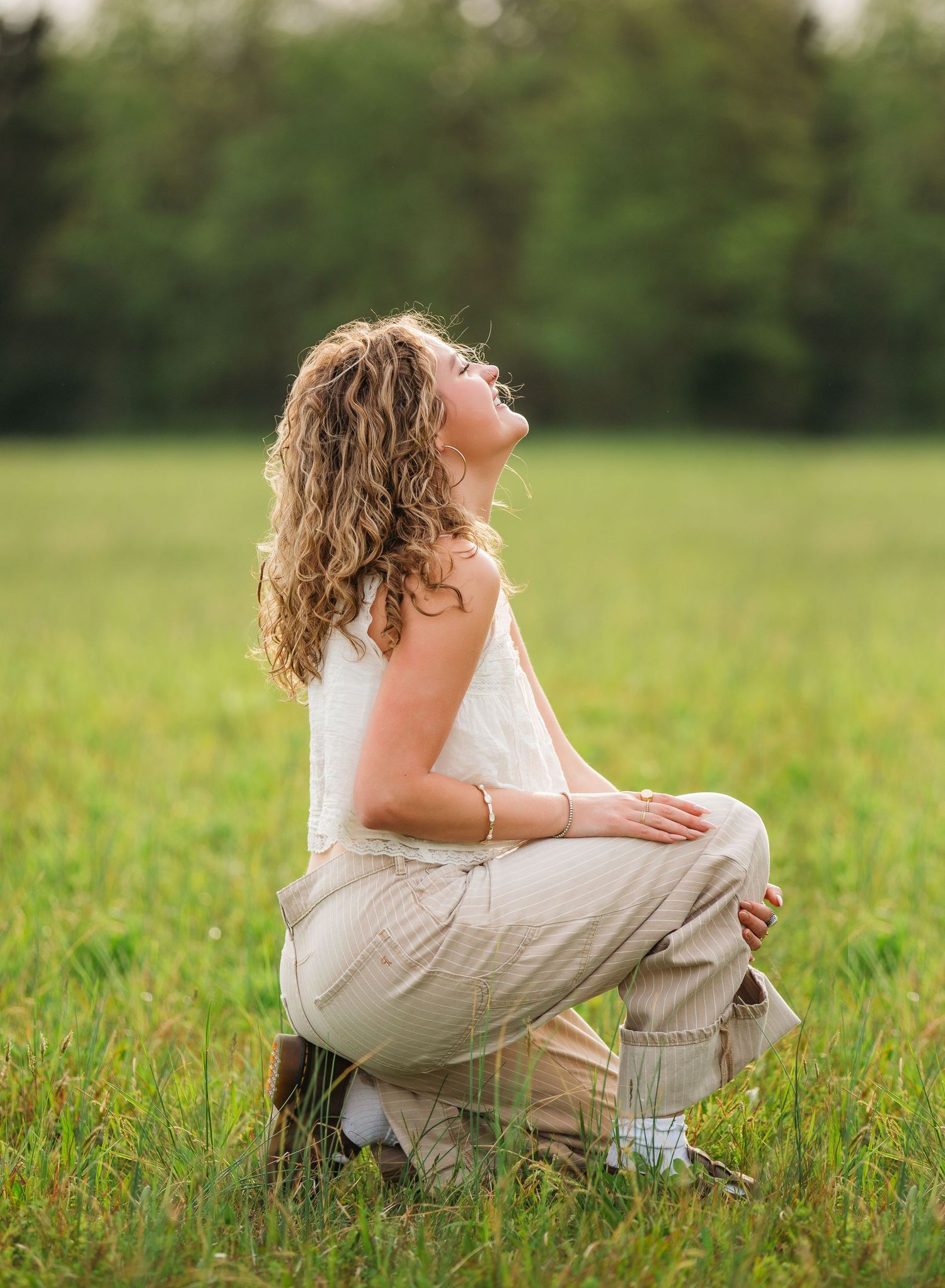 A person in a white top and khaki pants sits cross-legged in a grassy meadow, looking up at the sky with curly hair.