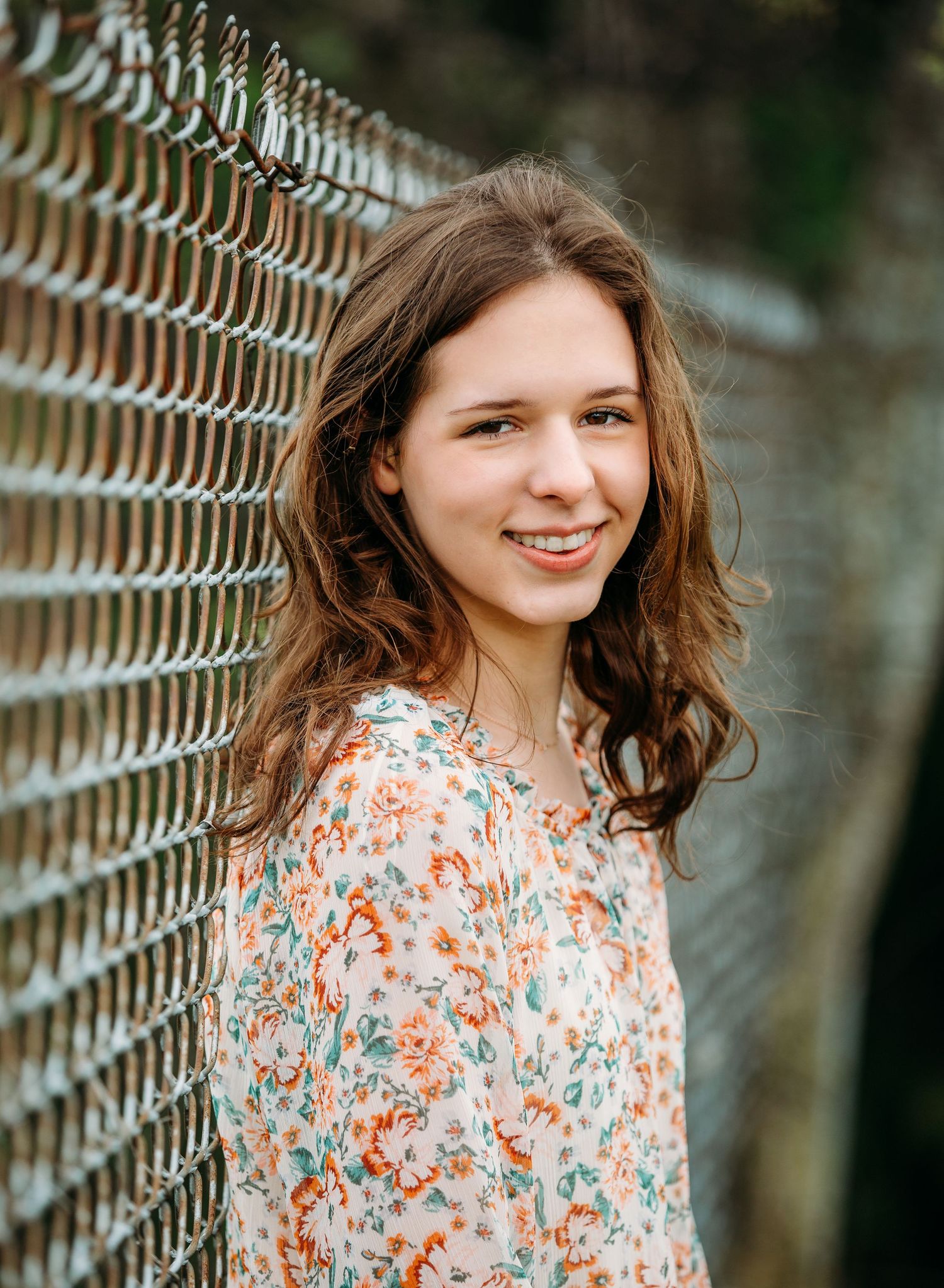 Someone with wavy brown hair and a floral print top smiles while leaning against a chain-link fence outdoors.