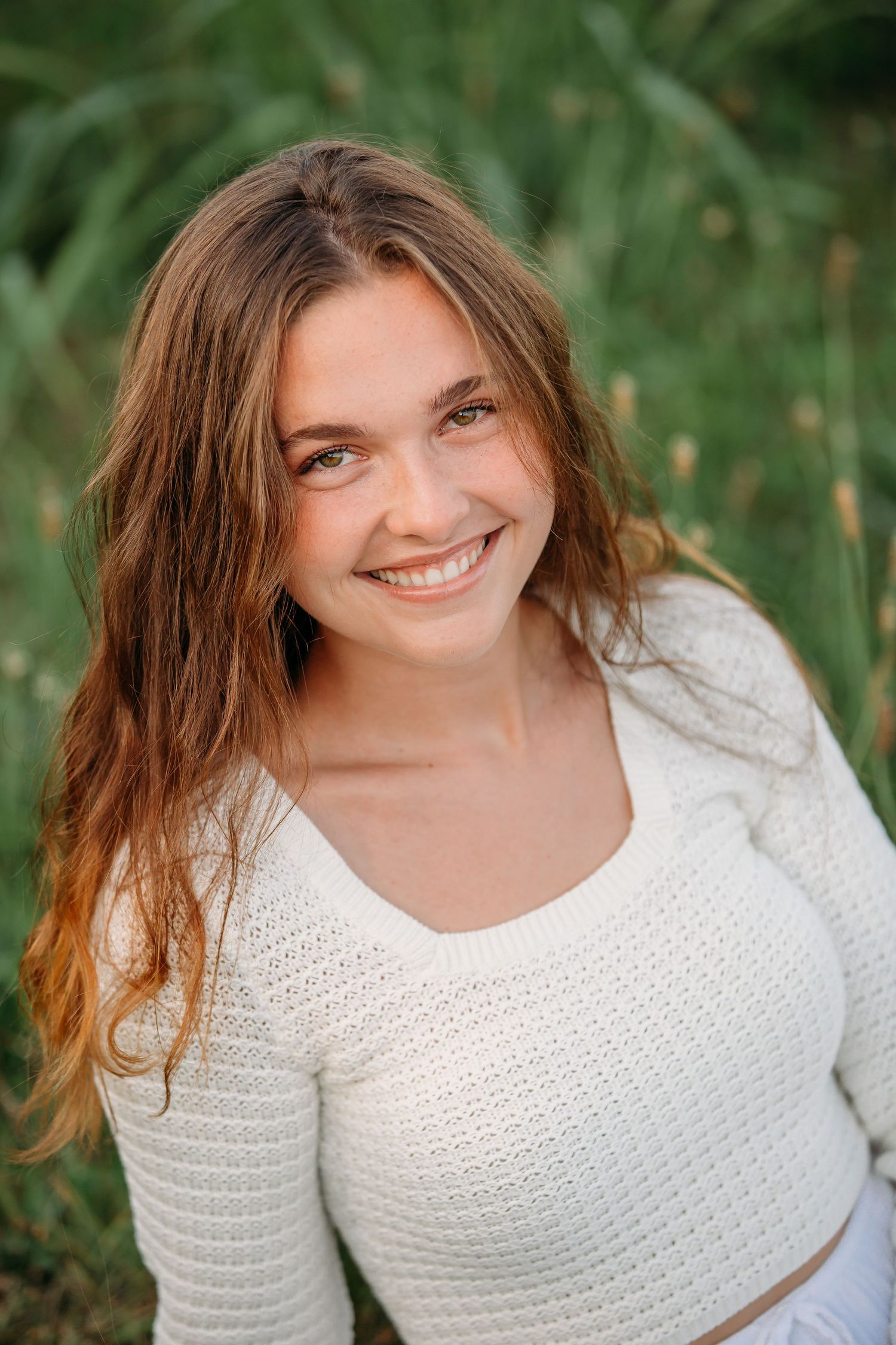 Portrait in natural outdoor setting wearing white knit sweater with long brown hair and genuine smile against green background.