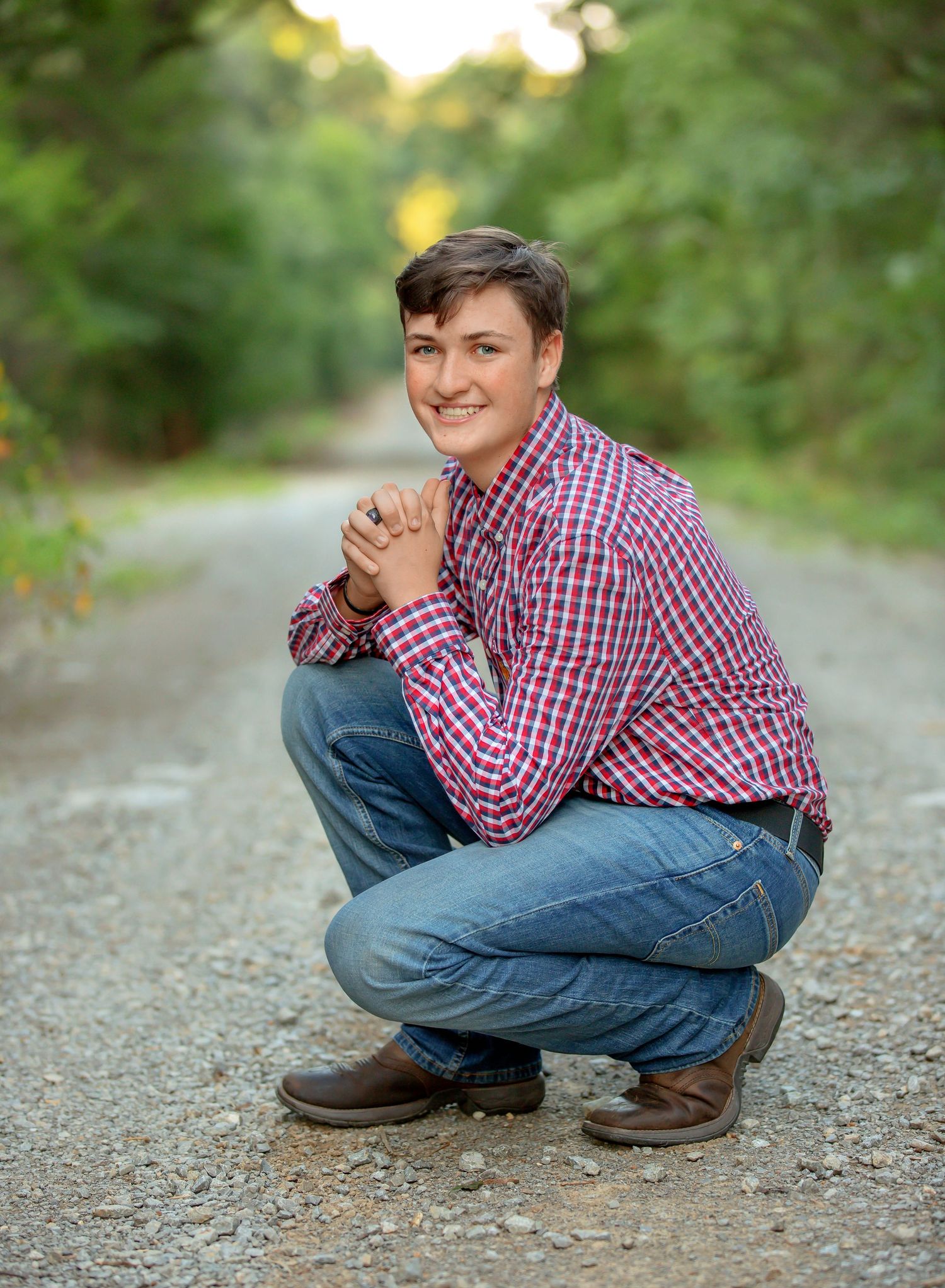 A person in a red plaid shirt and jeans crouches on a gravel path surrounded by lush greenery at sunset.