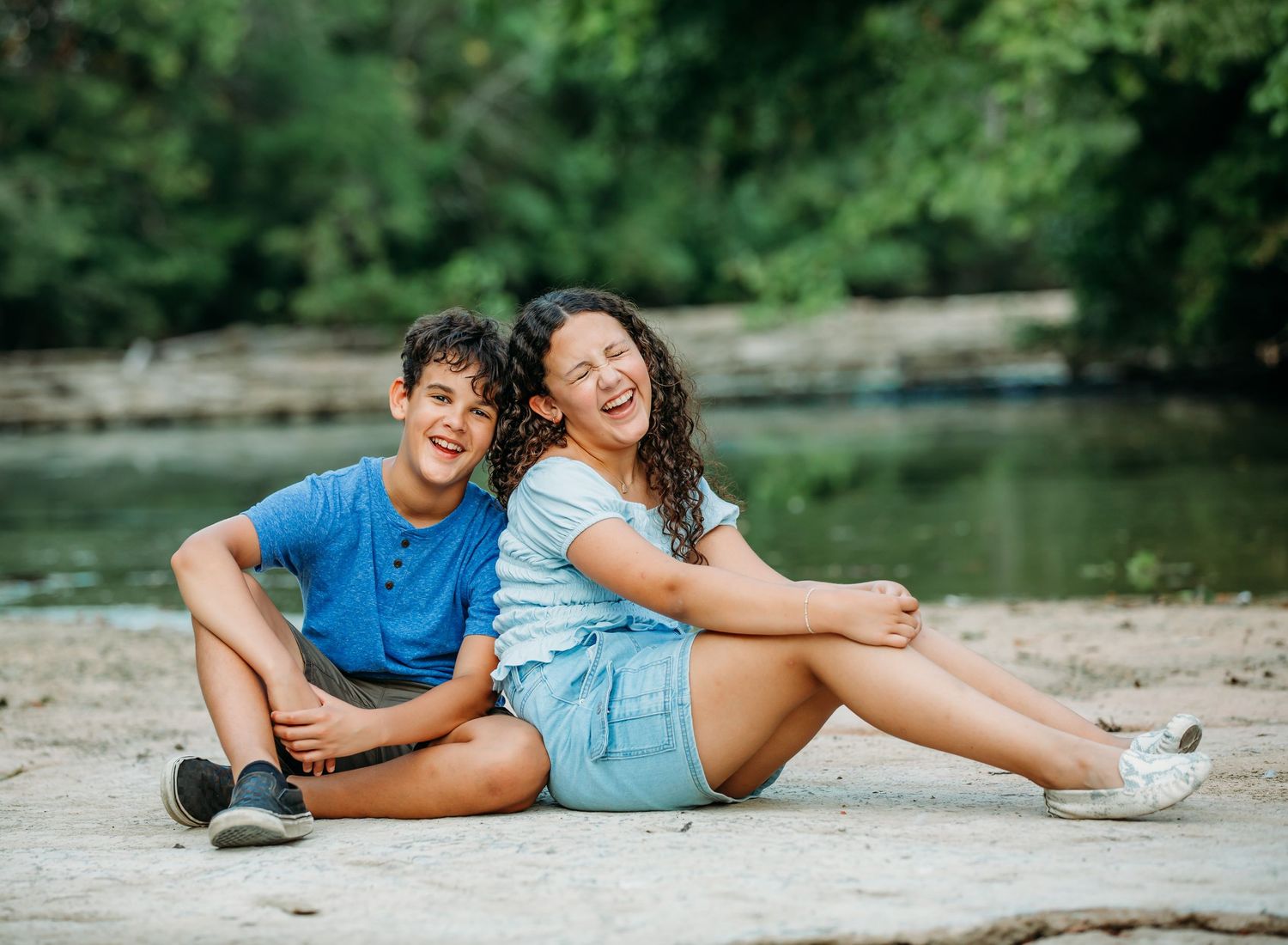 Two people sit together laughing on rocky shore by peaceful lake.