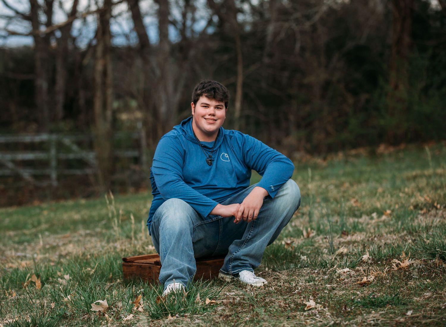 Person in casual denim attire sits on grassy slope during autumn with surrounding trees in background.
