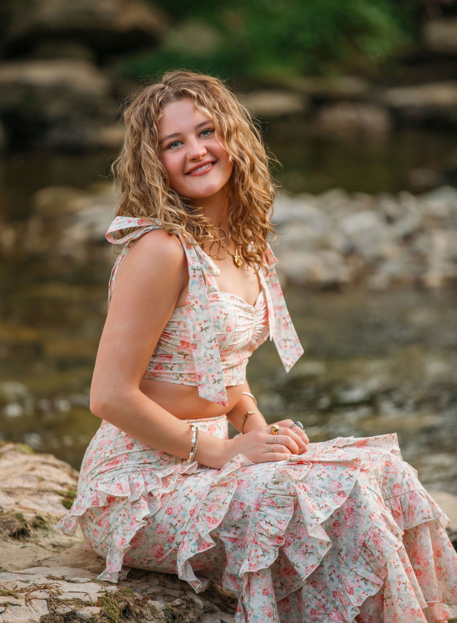 A person in a floral dress sits gracefully on rocks near a tranquil stream on a warm summer day.